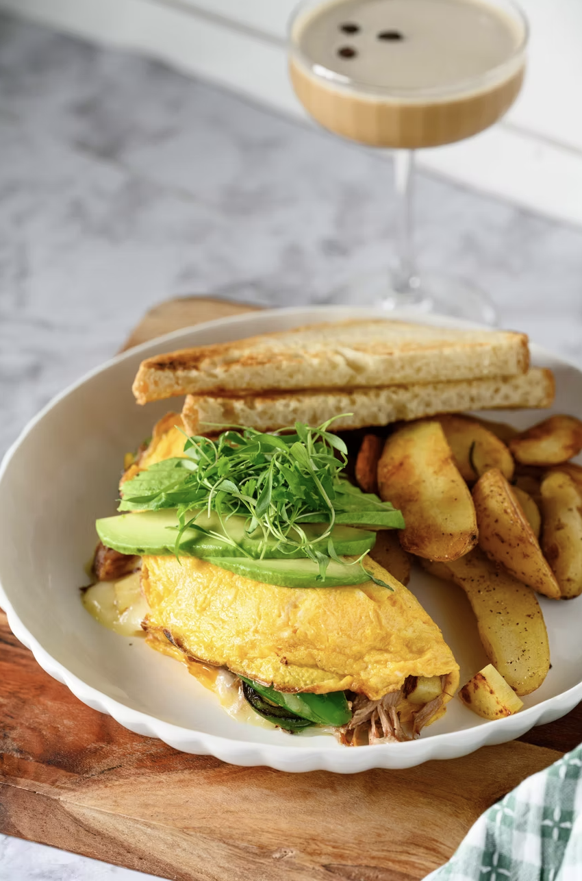 A plate with a breakfast sandwich, potato wedges, and fresh greens, with a latte in a glass on a gray marble surface.