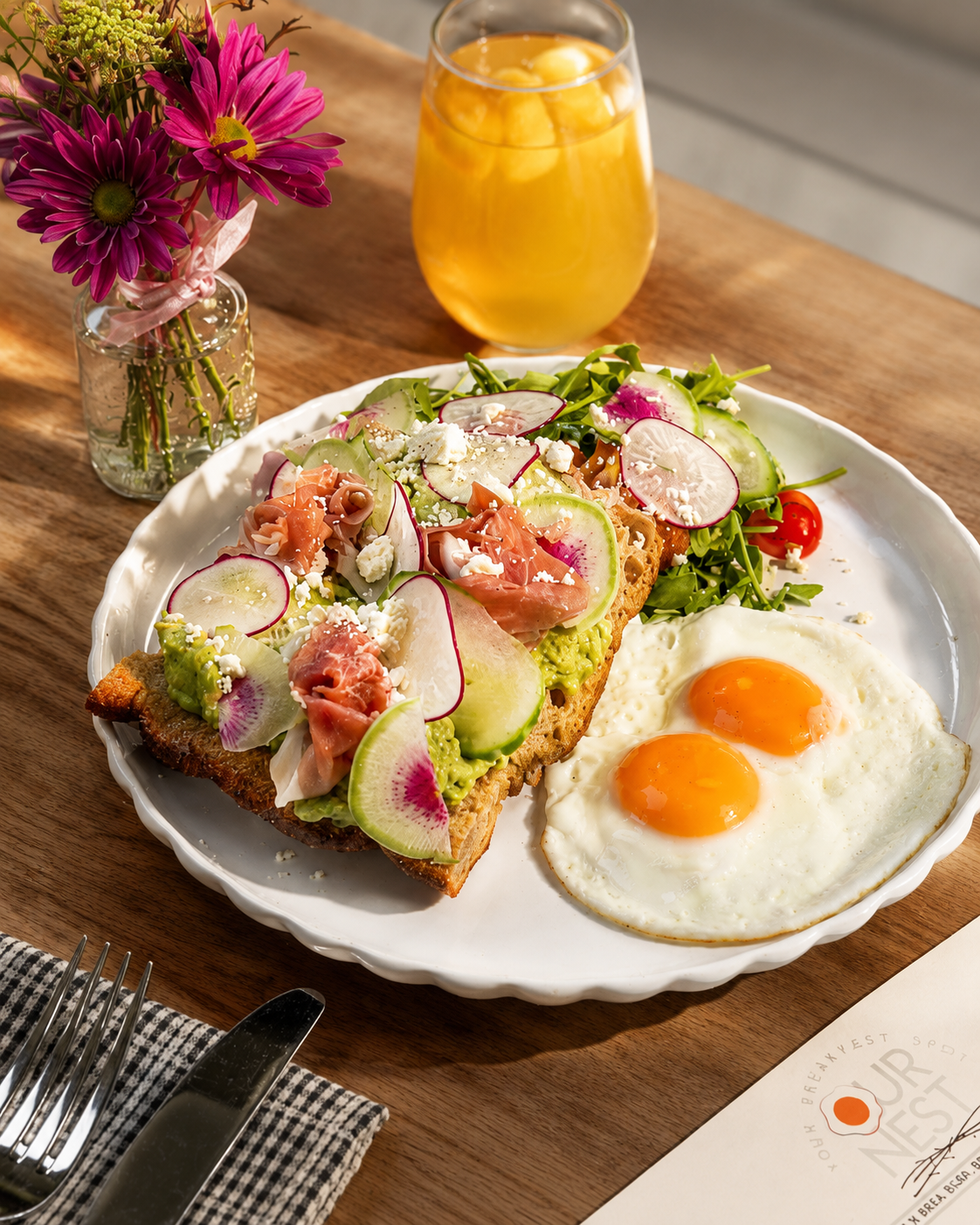 A breakfast plate with two sunny-side-up eggs, an avocado toast topped with radish slices, smoked salmon, and crumbled cheese, alongside a fresh mixed salad, on a wooden table with a glass of orange juice and a small vase of pink and purple flowers.