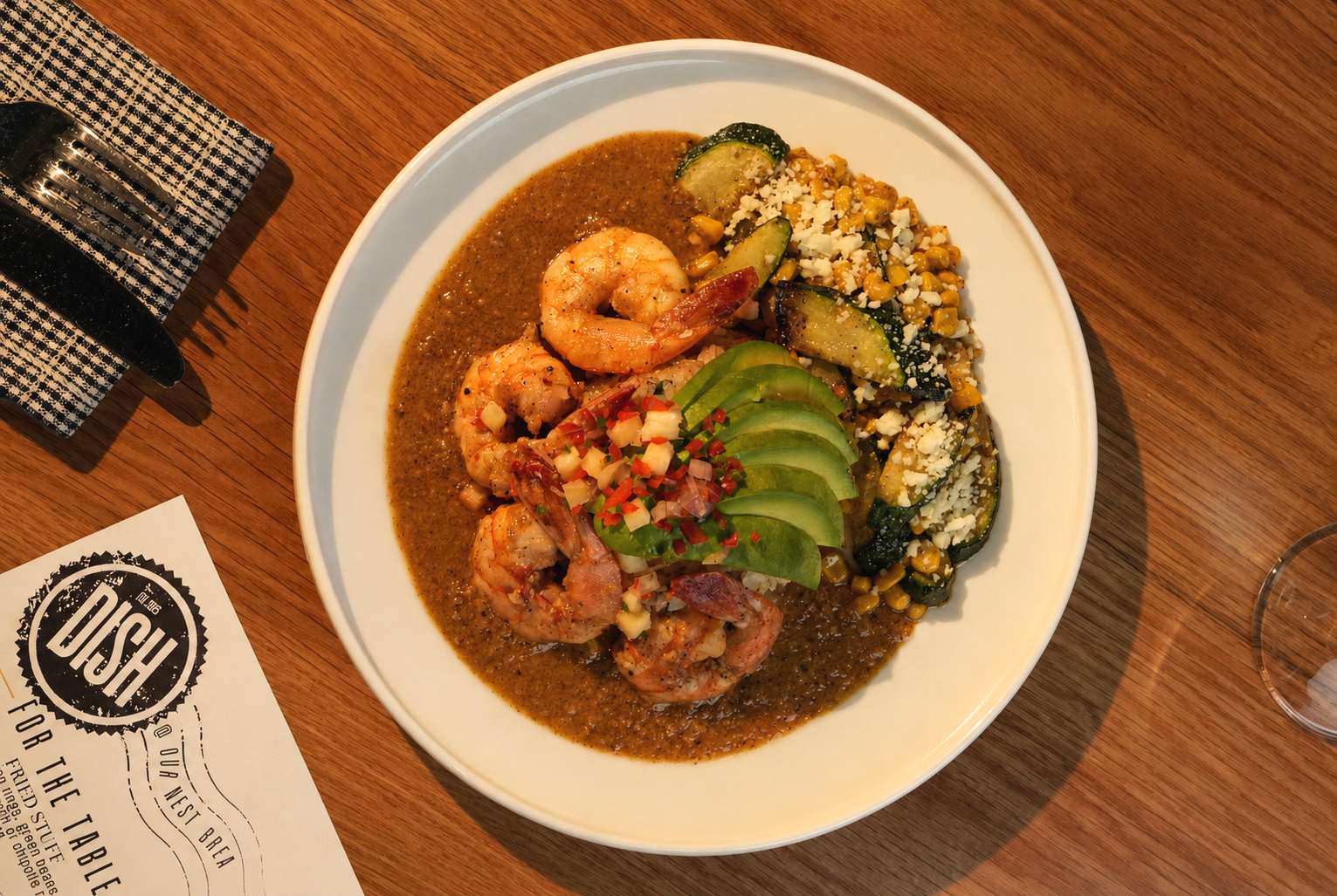 Seafood dish with shrimp and diced vegetables topped with avocado slices, served with a side of corn and zucchini, all on a white plate.