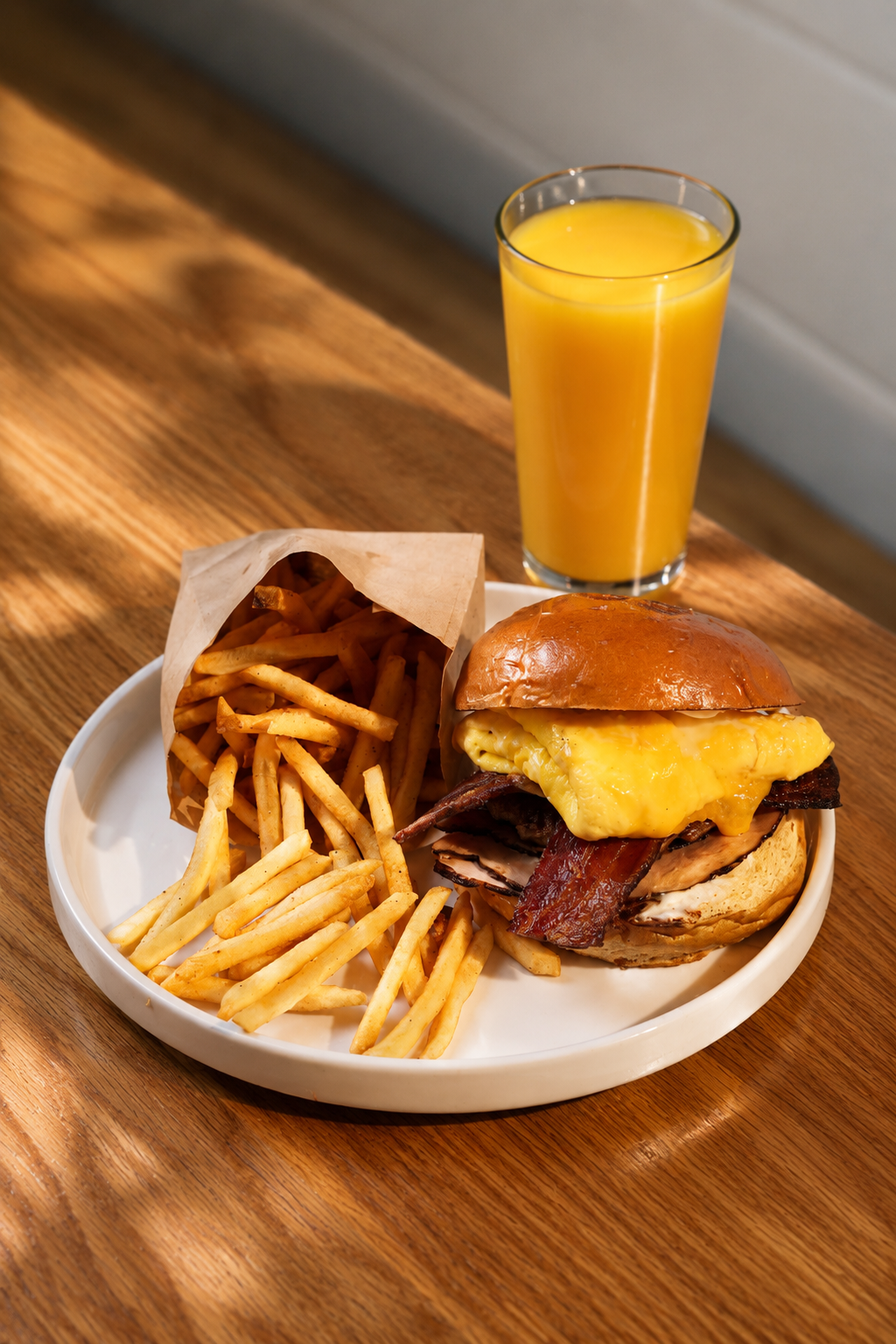 A plate with French fries and a breakfast sandwich containing bacon, sausage, egg, and cheese, alongside a glass of orange juice on a wooden table.