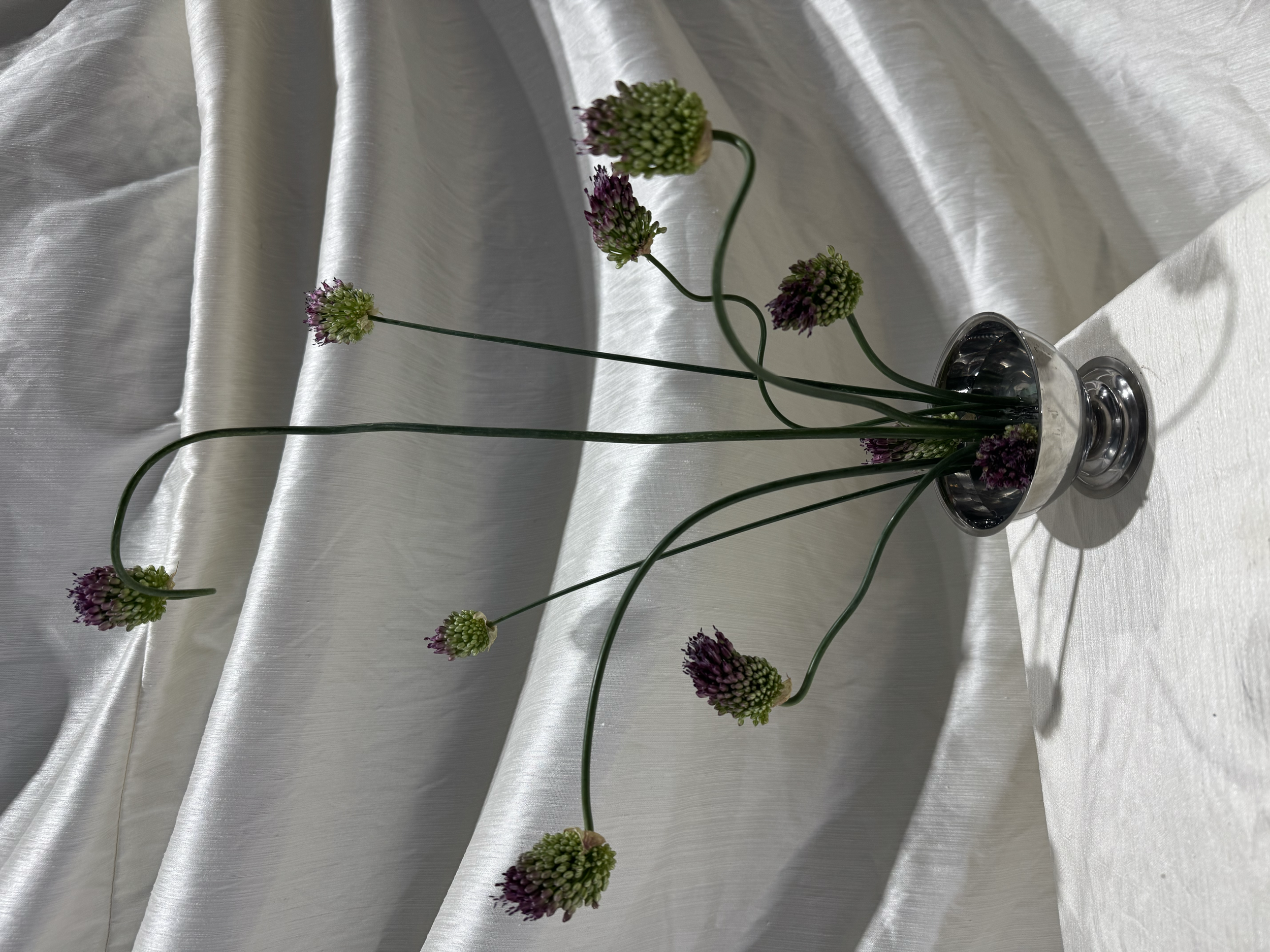 A silver vase with green and purple flowers placed on a white satin fabric background.