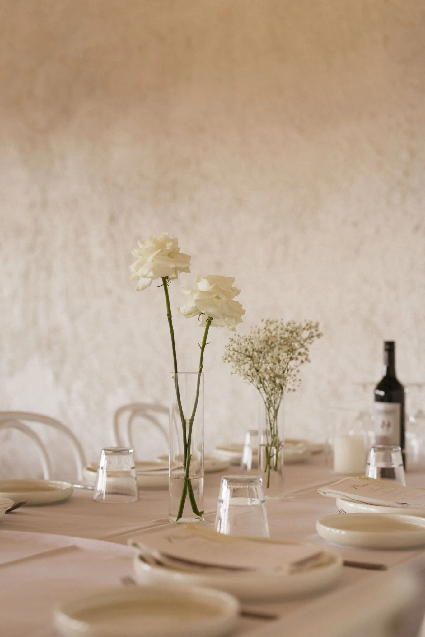 Elegant dining table setup with tall glass vases holding white flowers and smaller floral arrangements, set against a neutral wall background.