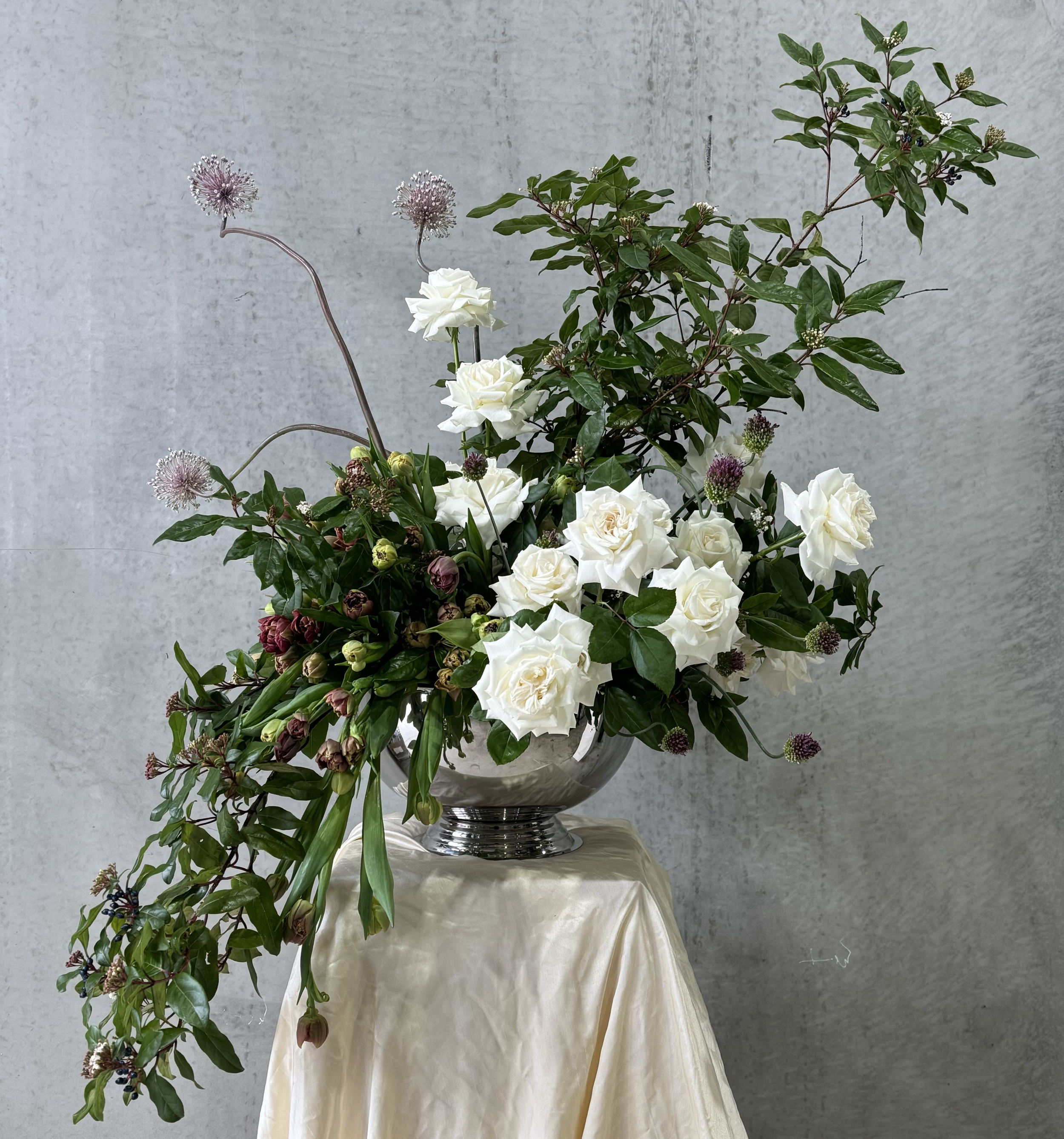 A bouquet of white roses and purple flowers in a silver vase, decorated with green leaves, set on a beige tablecloth against a gray wall background.