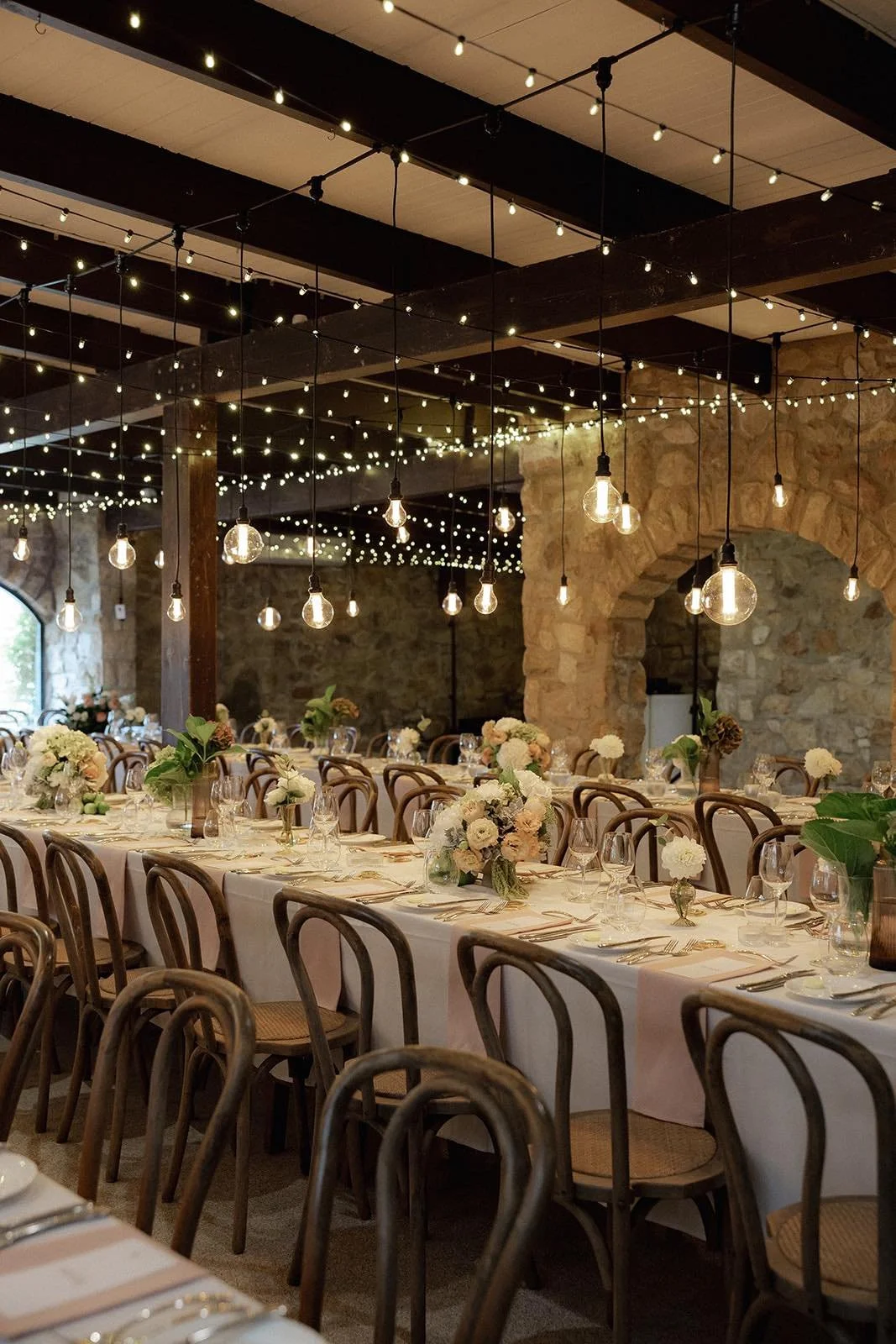 Wedding reception with long table set with white tablecloths, floral centerpieces, and glassware, decorated with string lights and bulbs hanging from the ceiling in a rustic stone venue.
