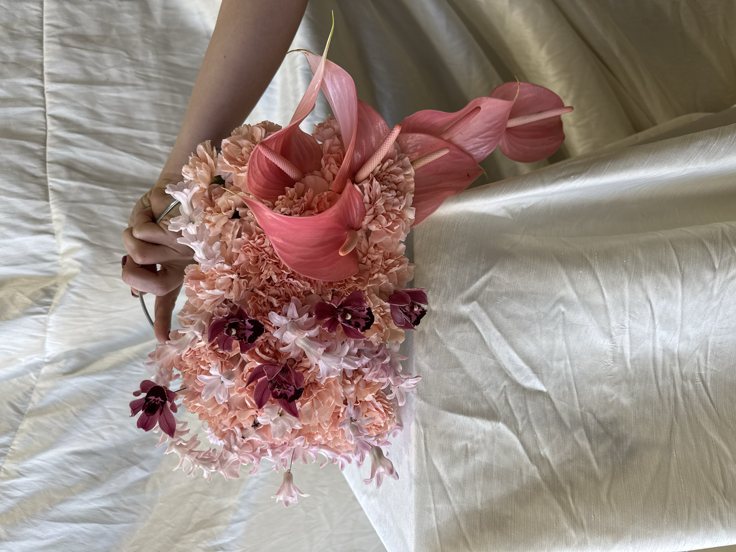 A floral arrangement with pink anthuriums, peach carnations, and purple orchids on a light-colored, satin-like fabric background.