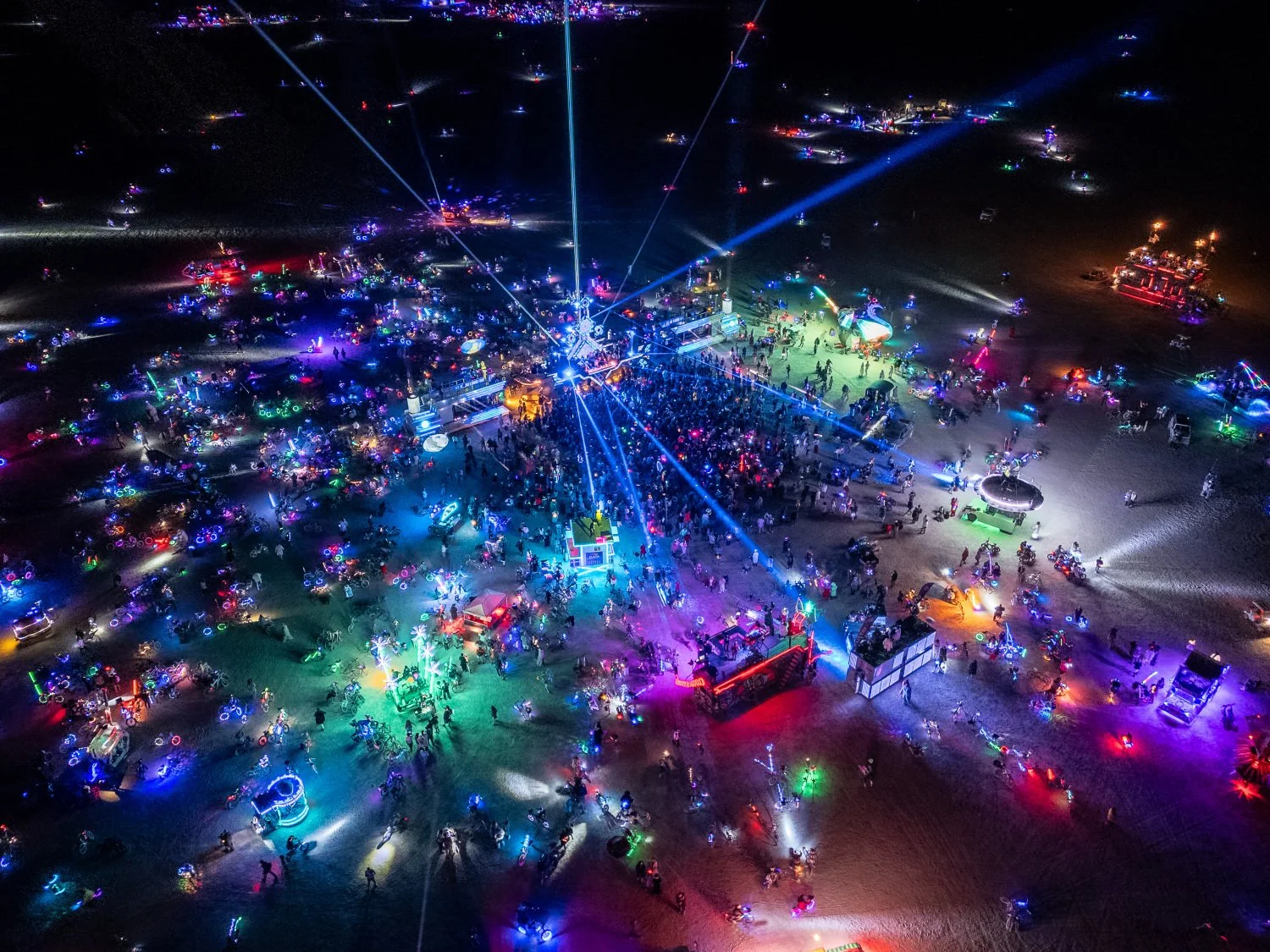 Vista aérea de un parque de diversiones nocturno con luces de neón y muchas personas disfrutando, con diferentes puestos y atracciones iluminadas.