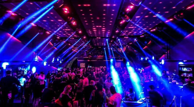 Interior de una discoteca con luces neón azules y rosas, y muchas personas bailando y socializando.
