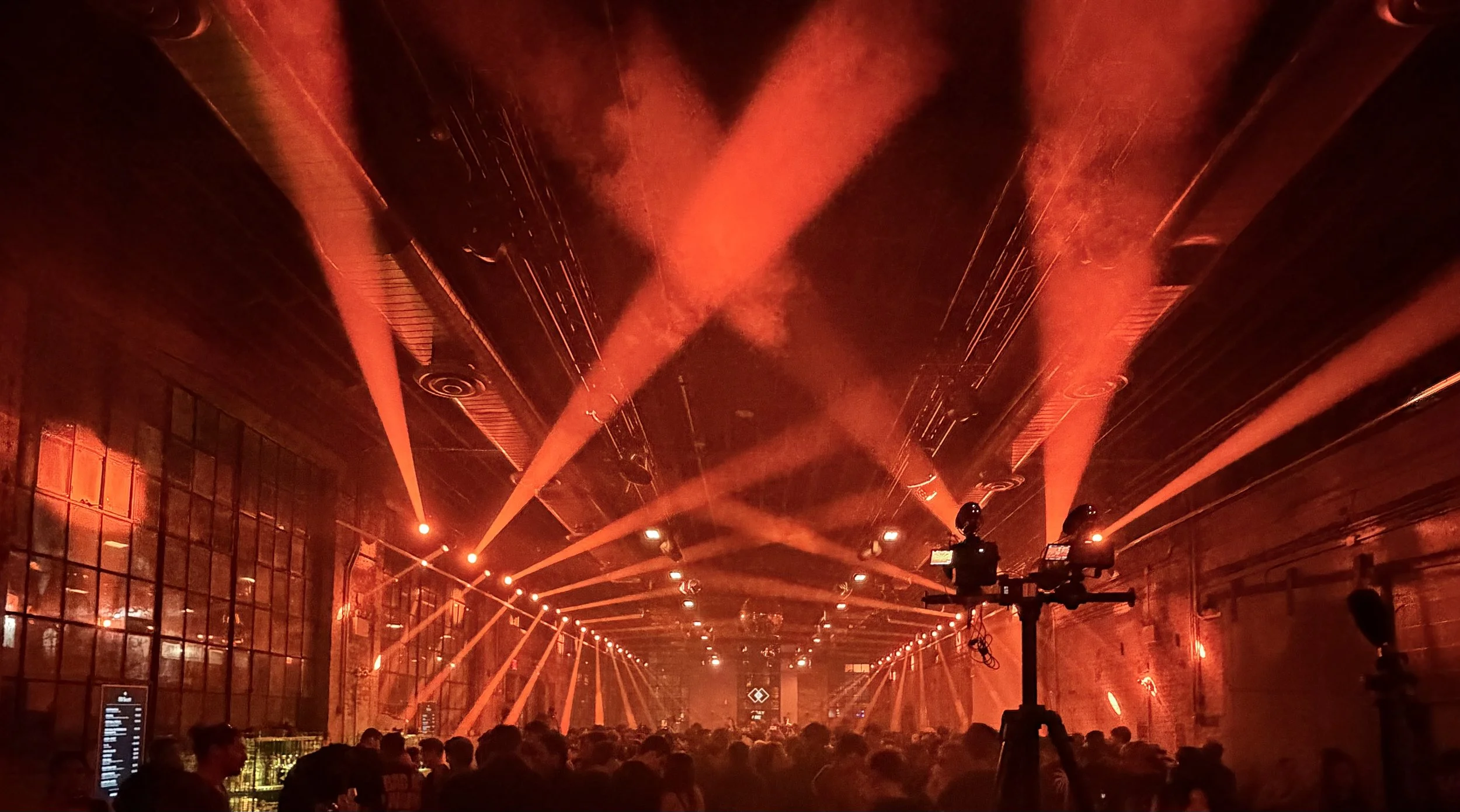 Una vista del interior de un concierto o evento con iluminación roja intensa y efectos de humo en el techo, con muchas personas en el público y algunos equipos de DJ o sonido en el escenario.