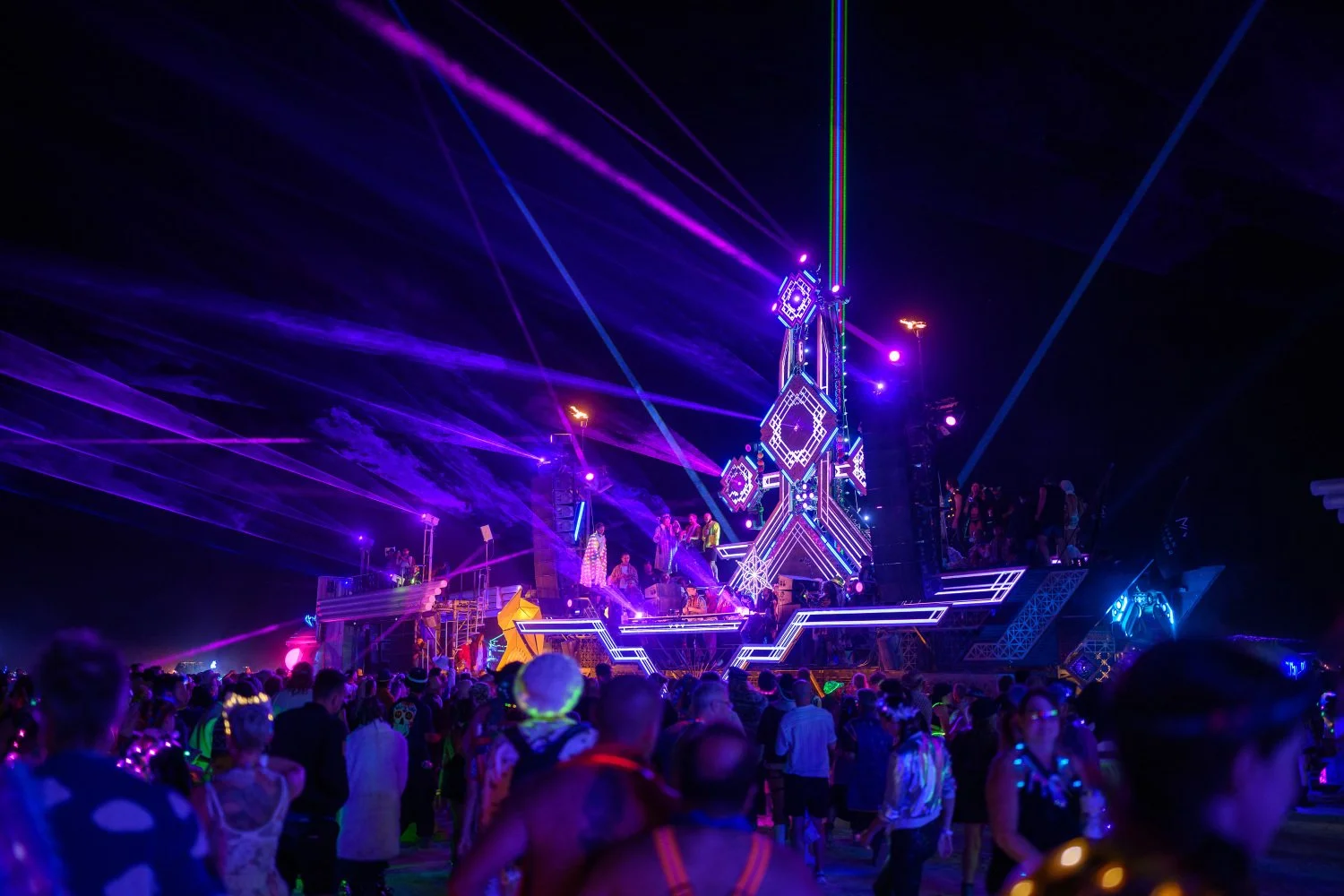 Escenario de un festival de música con luces de colores y láseres en la noche, lleno de personas disfrutando del evento.