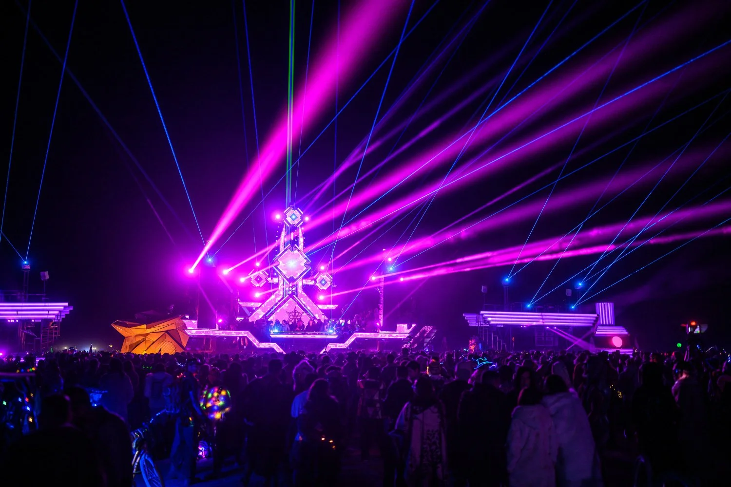 Escenario de concierto con luces láser rosas y azules, con una multitud de personas disfrutando la música en la noche.