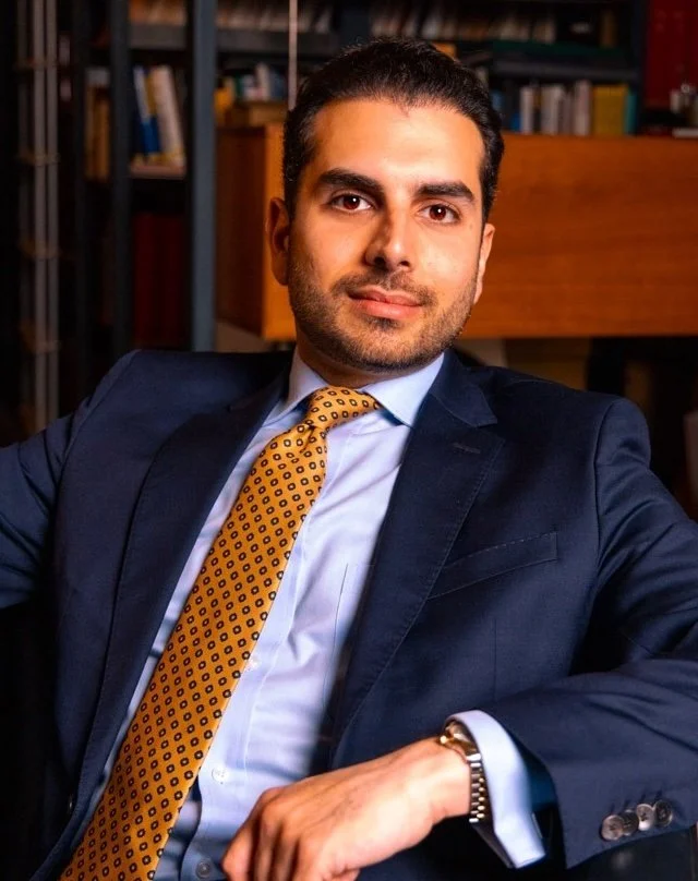 Ali Maisam Nazary in a dark suit with a yellow patterned tie sits in a professional office environment with a bookshelf in the background.