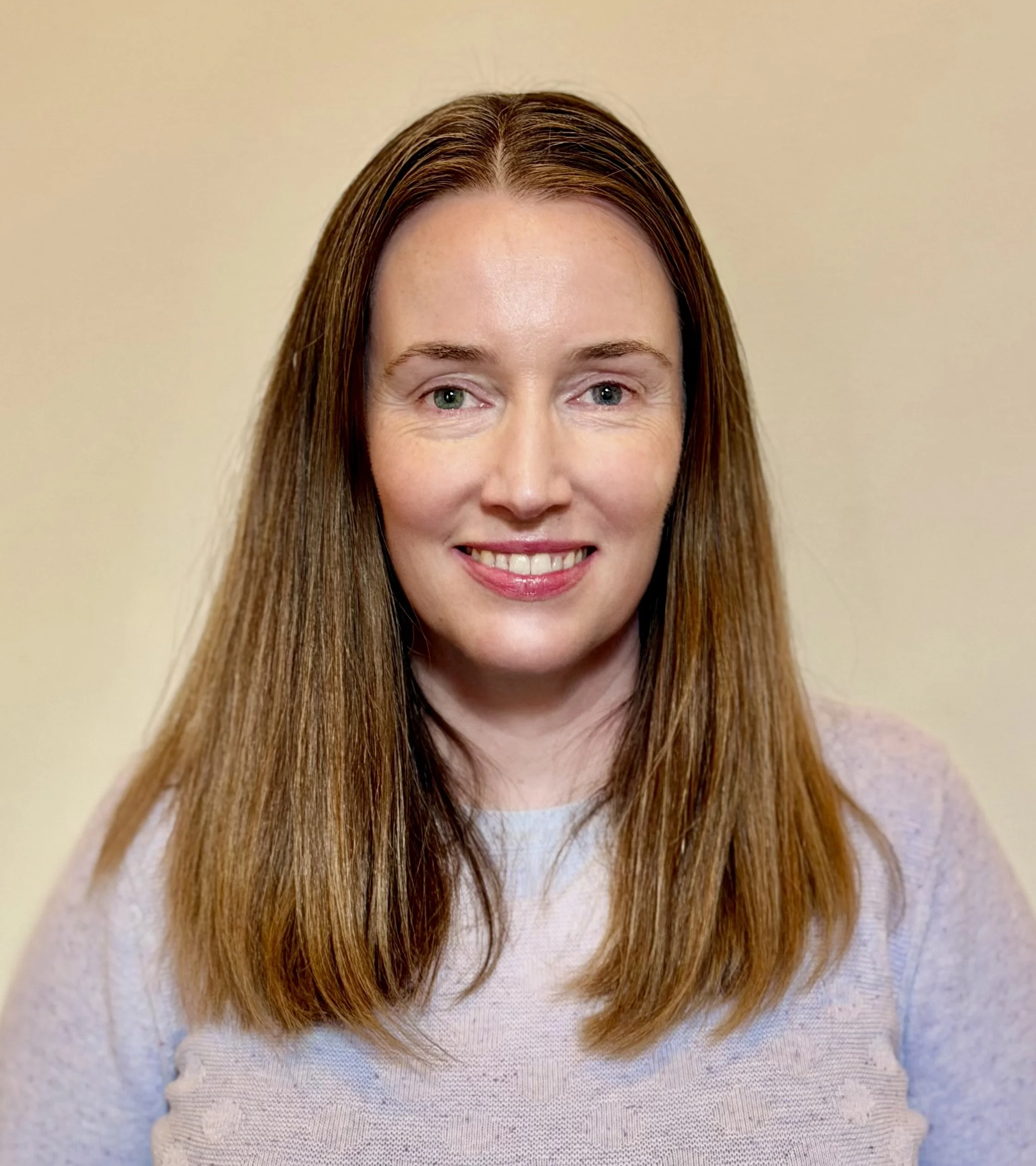 Close-up of a woman with straight, reddish-brown hair, smiling, wearing a light-colored, textured sweater, against a neutral background.