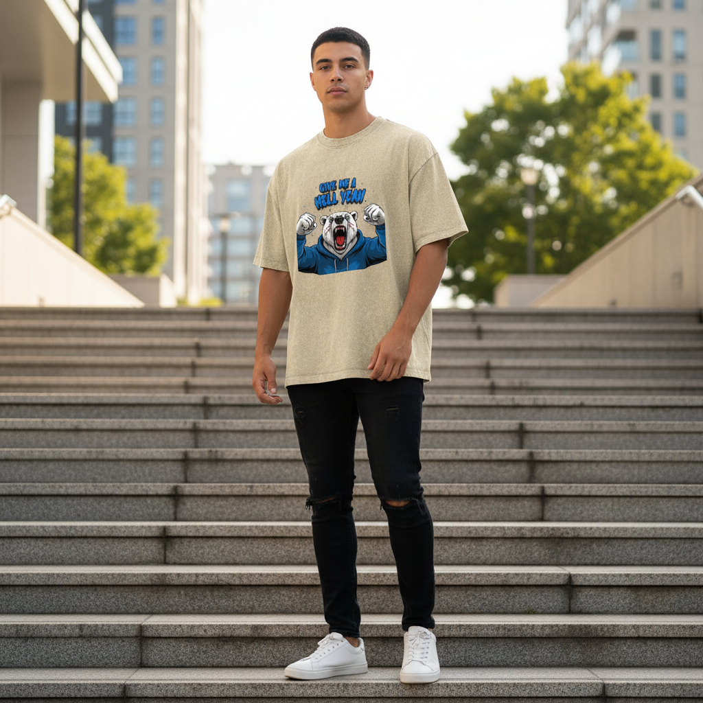 A young man standing on outdoor stairs in an urban setting, wearing a beige oversized T-shirt with a graphic of a roaring bear and the text 'Give Me a Hell Yeah,' black ripped jeans, and white sneakers.