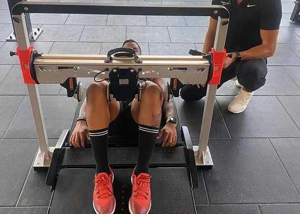 Junge Person liegt auf einer Beinpressen-Maschine im Fitnessstudio, betreut von einem Trainer