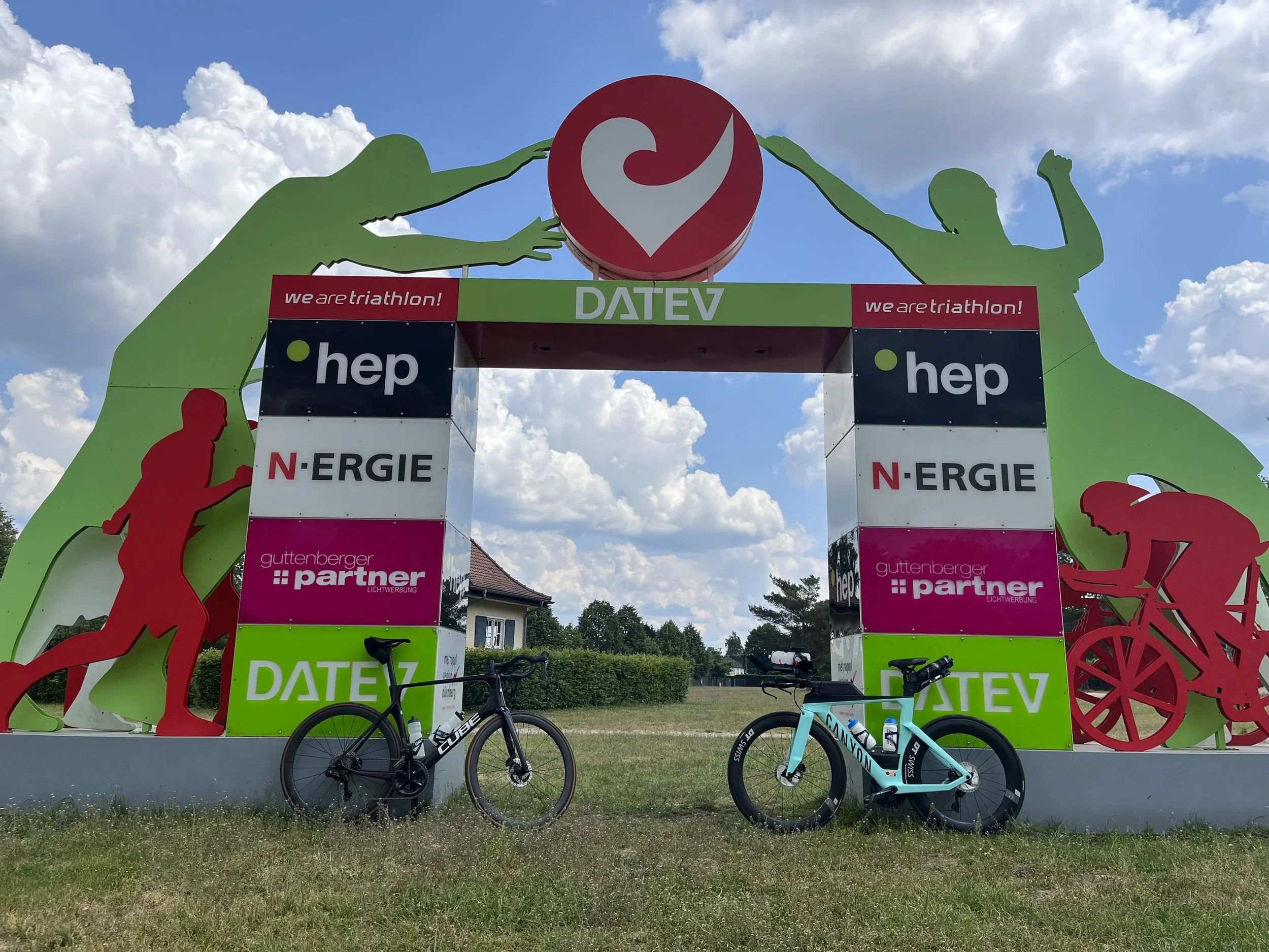 Bühne mit Sportmotiven, darunter Radfahrer-Silhouetten, und Beschriftungen wie „we are triathlon!“, „N-ERGIE“, „gutenberger partner“ und „DATZ“. Fahrräder sind im Vordergrund abgestellt.