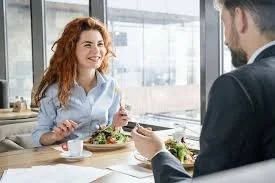 Zwei Menschen, eine Frau mit roten Locken und ein Mann, sitzen sich beim Essen in einem modernen Café oder Restaurant gegenüber. Sie sprechen und lachen.
