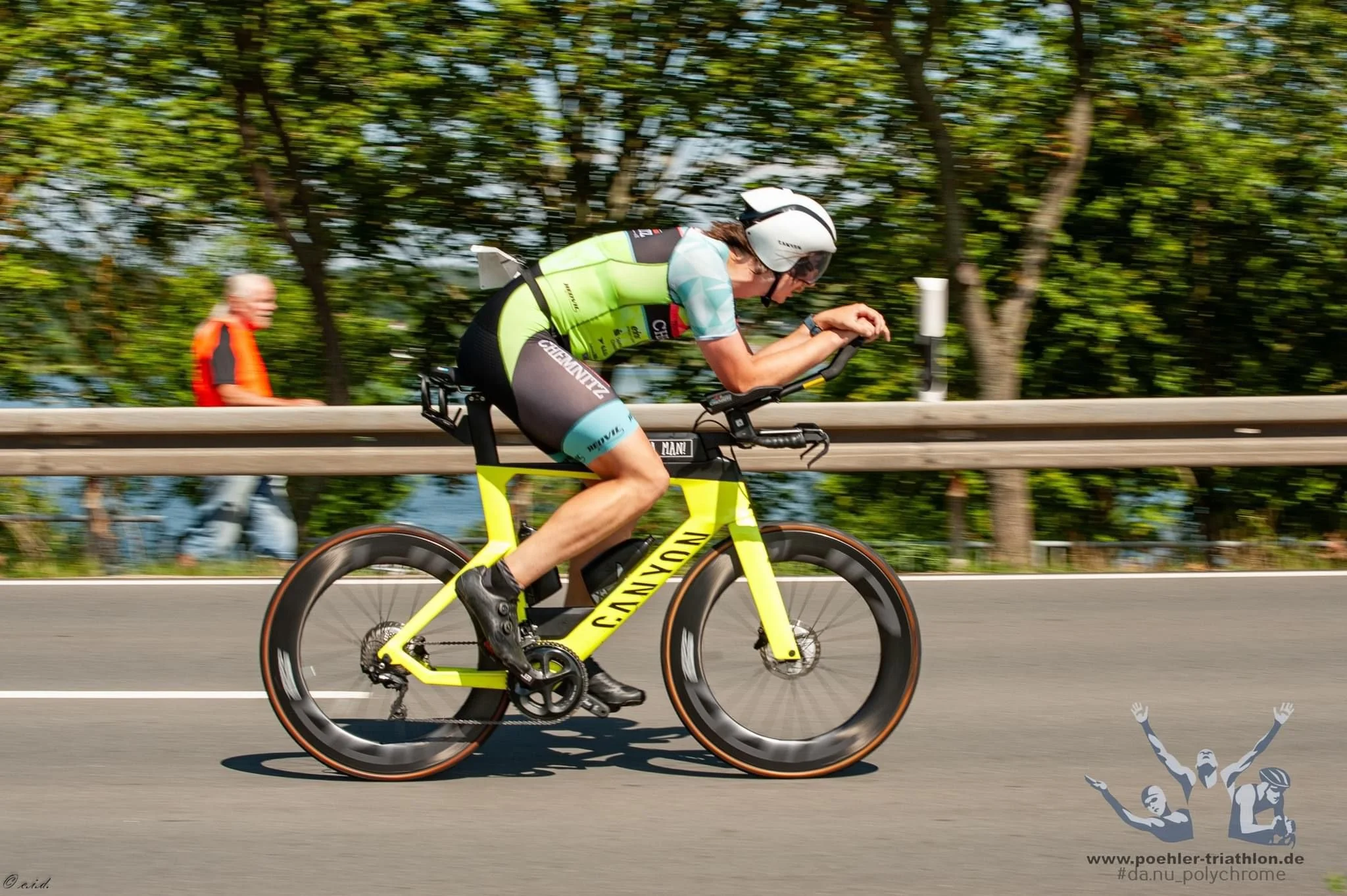 Ein Radfahrer in neonfarbener Fahrradkleidung und Helm fährt auf einer Straße, im Hintergrund sind Bäume. Im unteren rechten Ecke befindet sich ein Logo mit einer sportlichen Figur und Text. Es ist eine Actionaufnahme mit Bewegungsunschärfe.