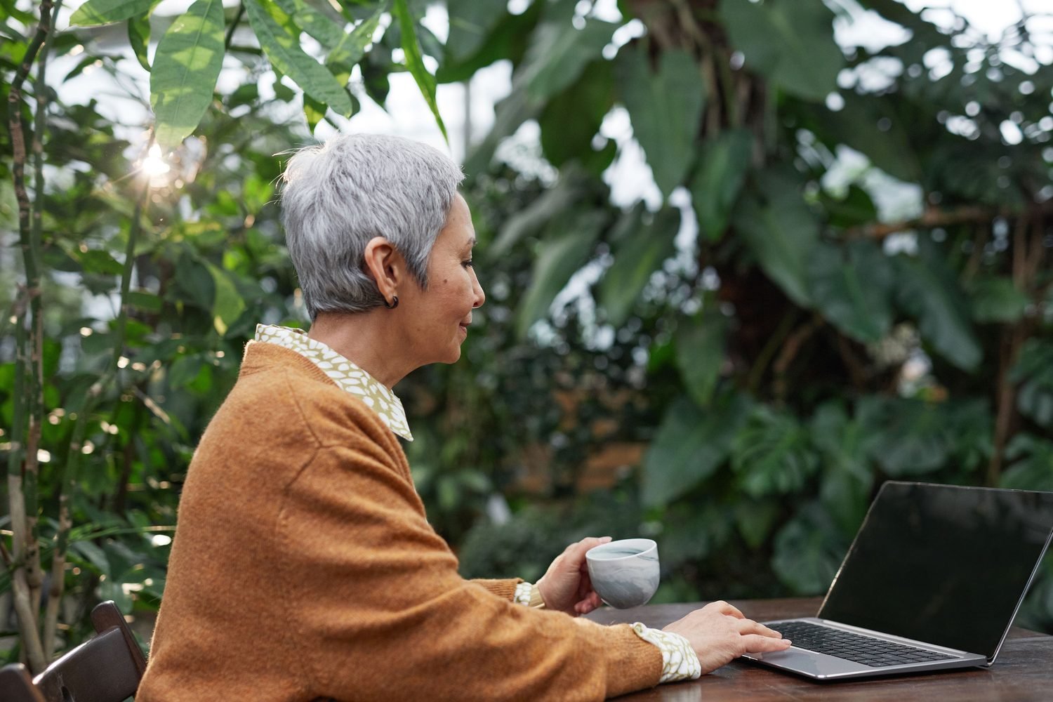 Woman sipping tea while working on her laptop outdoors, surrounded by greenery