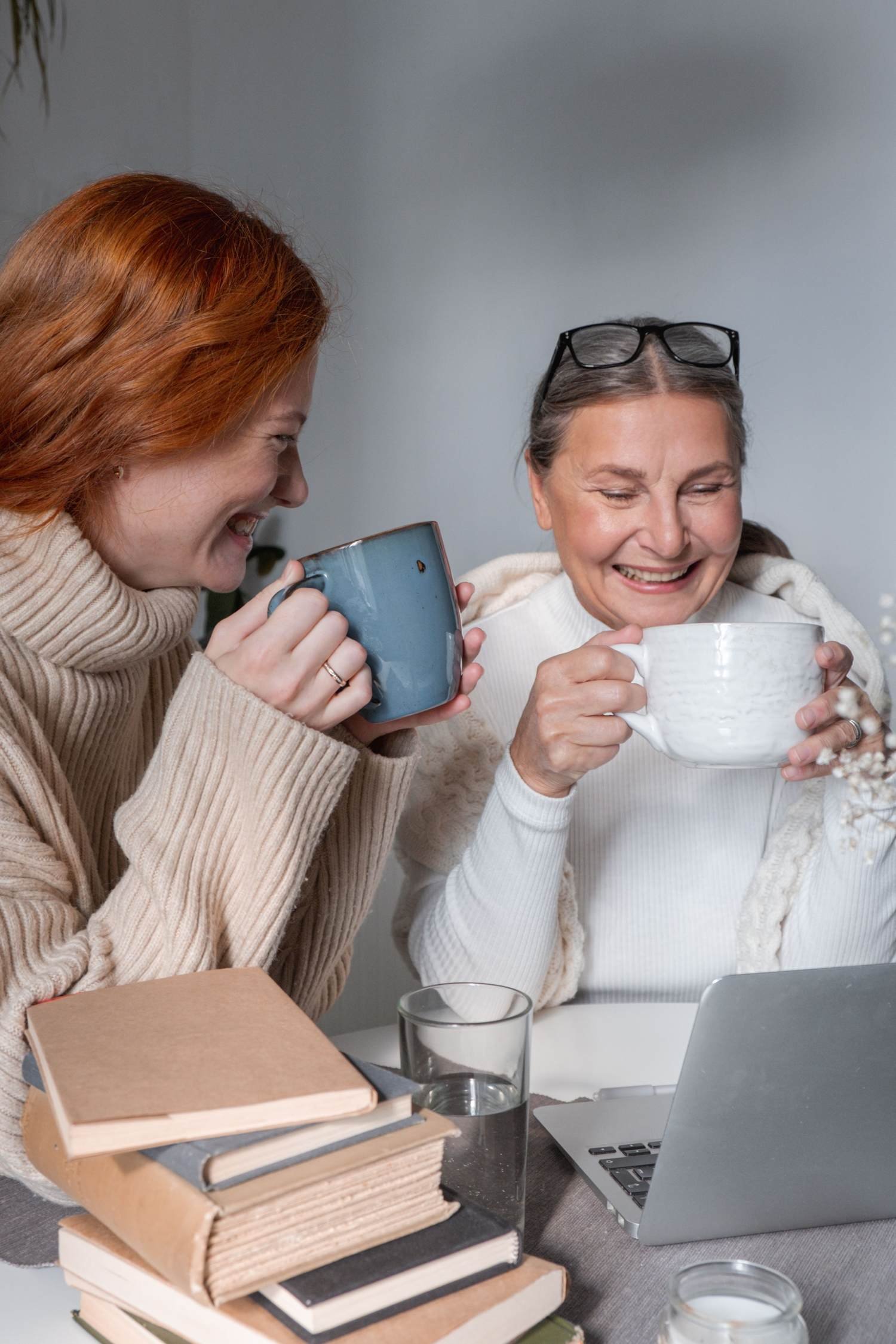 Two women laughing together over coffee, books stacked nearby