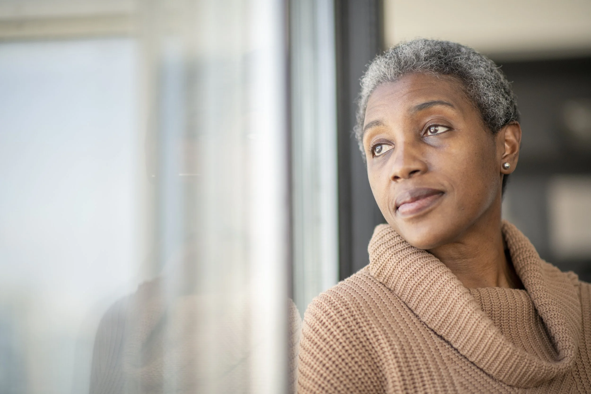 Woman in her fifties looking out a window, reflecting on sudden crashing fatigue at midlife.