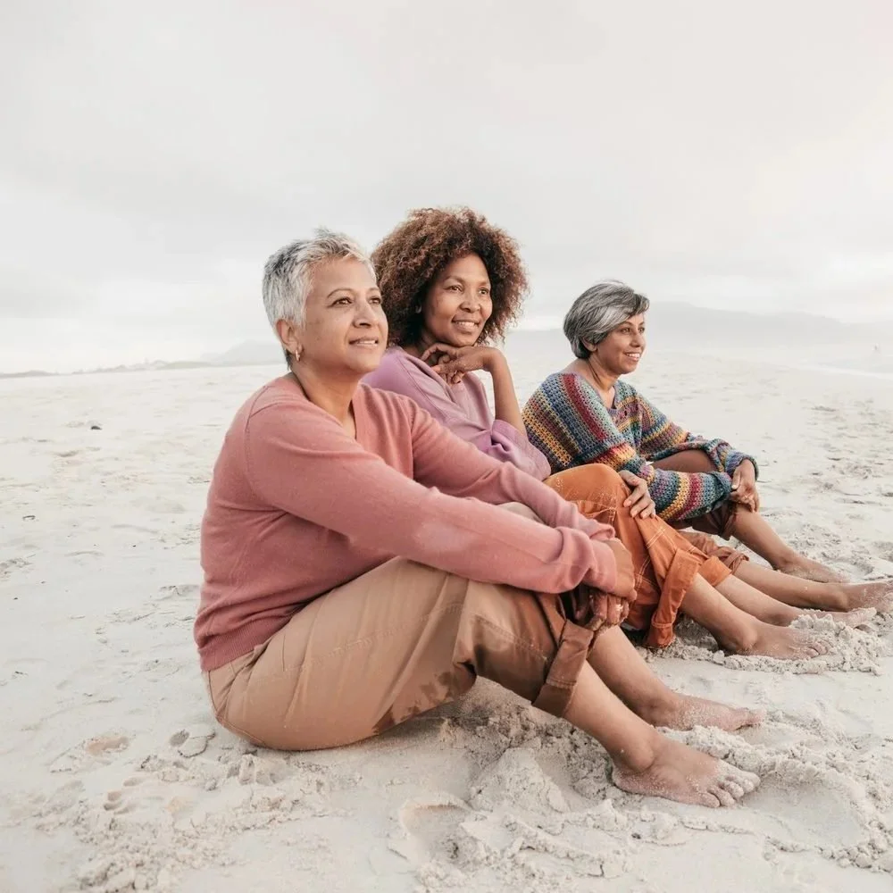 Three women sitting together on the beach, laughing and enjoying each other's company