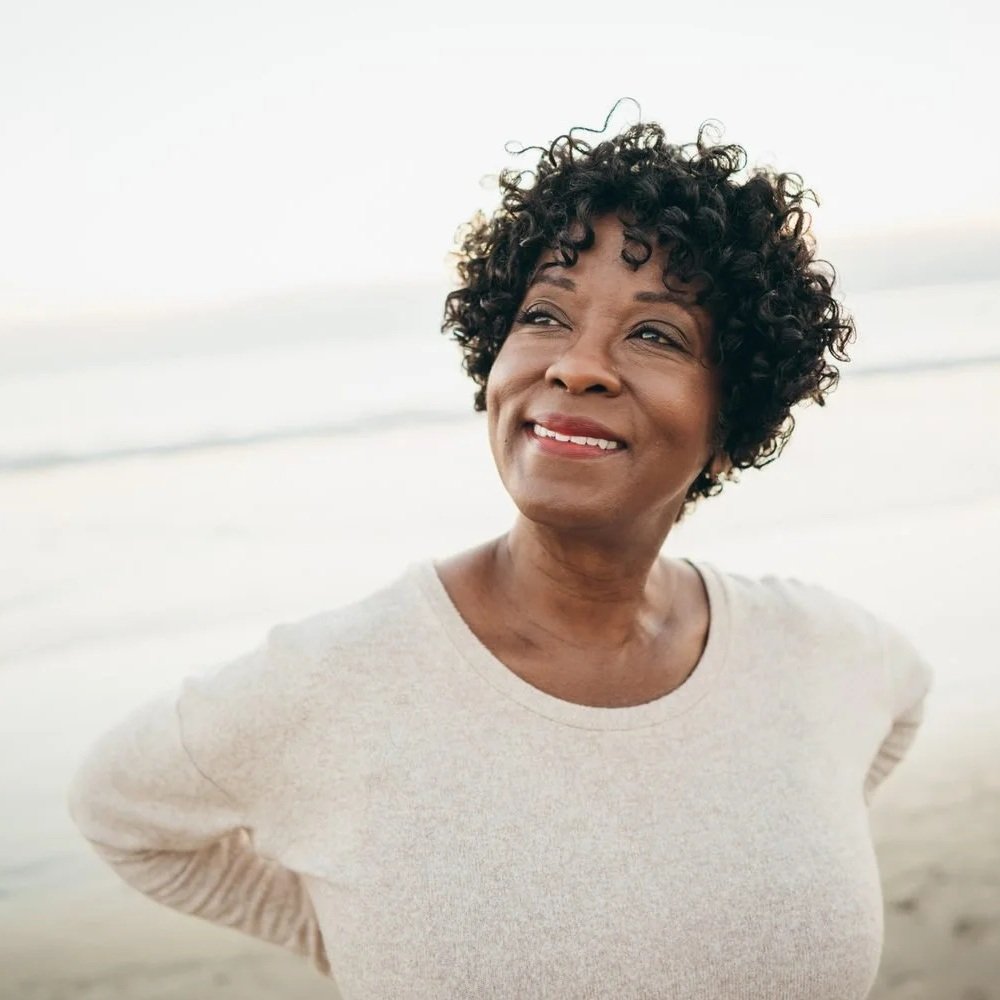 Smiling woman with short curly hair standing on the beach, embodying quiet confidence