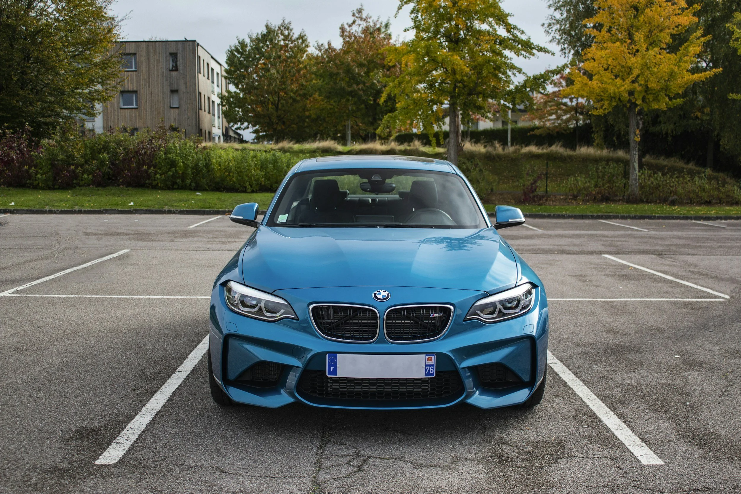 A bright blue BMW car parked in an empty parking lot with white parking lines, surrounded by trees with autumn-colored leaves and a multi-story building in the background.