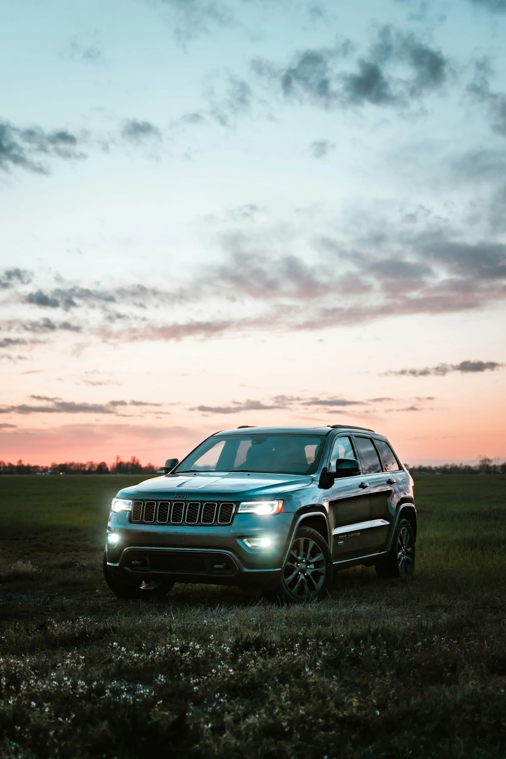 A dark gray Jeep SUV parked on a grassy field during sunset, with a colorful sky filled with clouds and the horizon in the background.