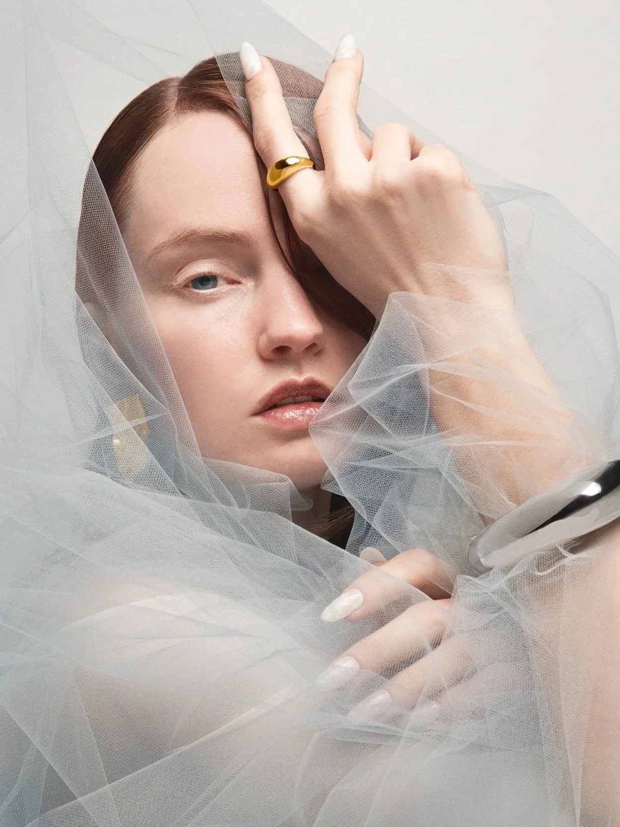 A close-up of a woman with fair skin and light blue eyes, surrounded by sheer white tulle fabric, holding the fabric with one hand and making a peace sign near her face.