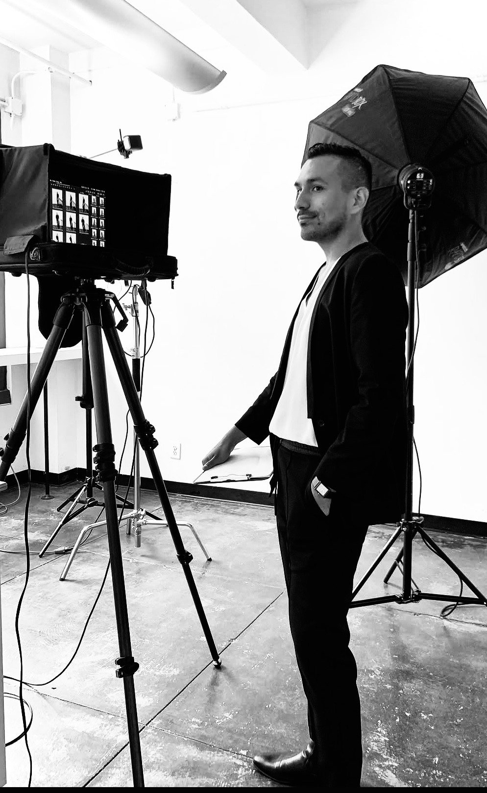 A man in a suit and white shirt standing in a photography studio with a camera on a tripod, softbox light, and a backdrop. The image is in black and white.