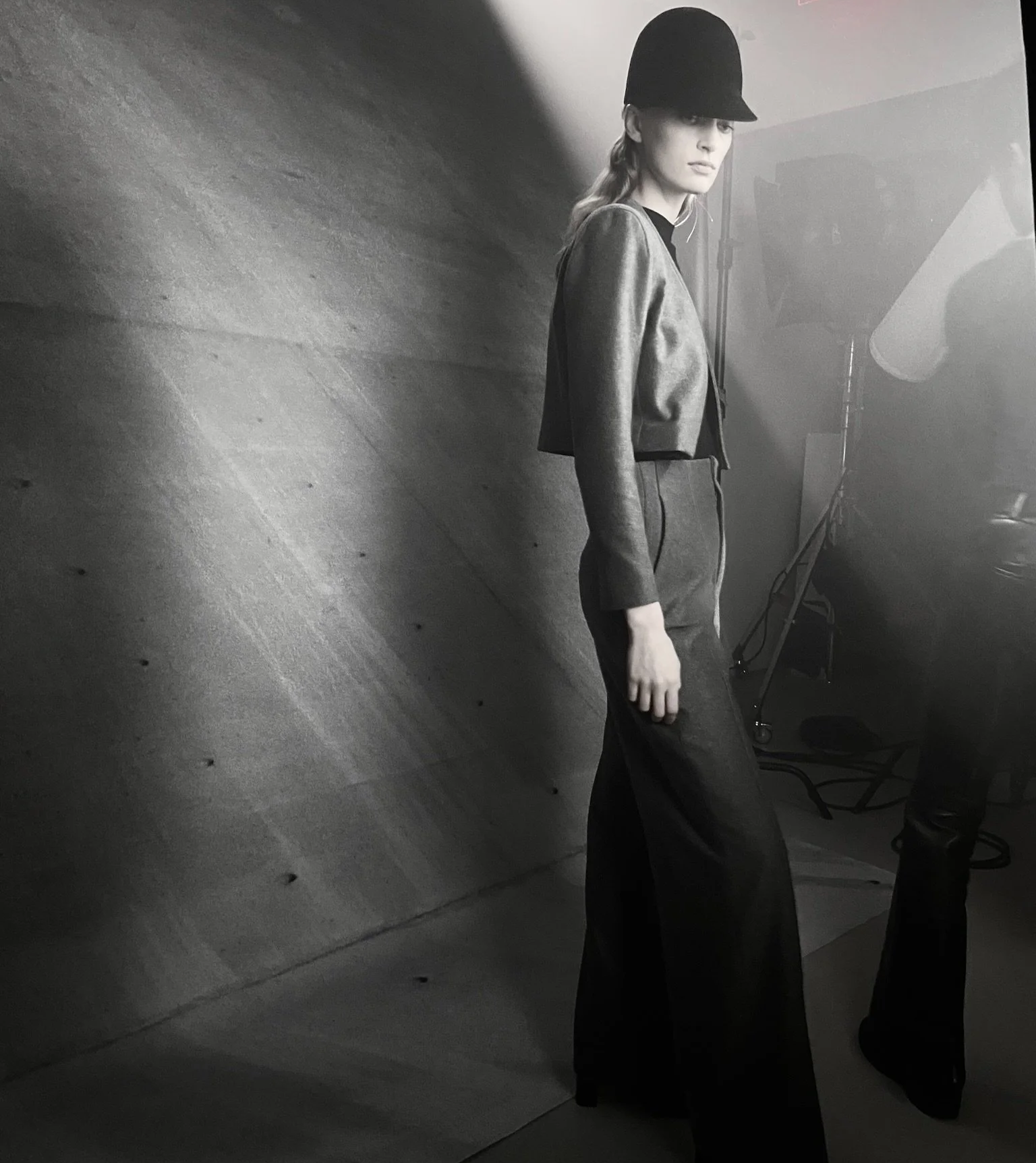 Black and white photo of a woman with light hair, wearing a hat and a cropped jacket, standing against a textured wall. She is looking directly at the camera with a serious expression.