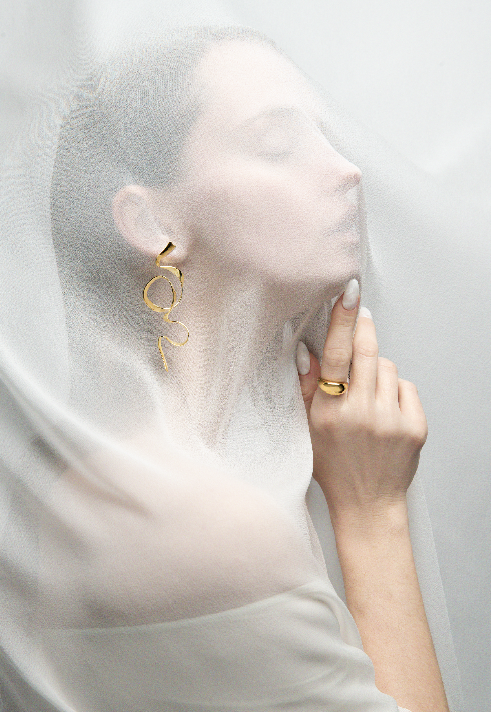 A woman with closed eyes and minimal makeup, wearing large gold earrings and a gold ring, touches a semi-transparent fabric or veil in front of her face, creating a soft, ethereal effect.