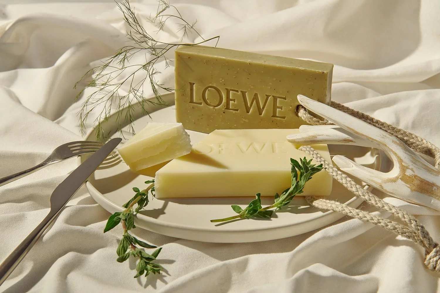 Various bars of soap with the brand name "LOEWE" embossed on them, arranged on a white plate with some greenery and a small tool on a cream-colored fabric background.