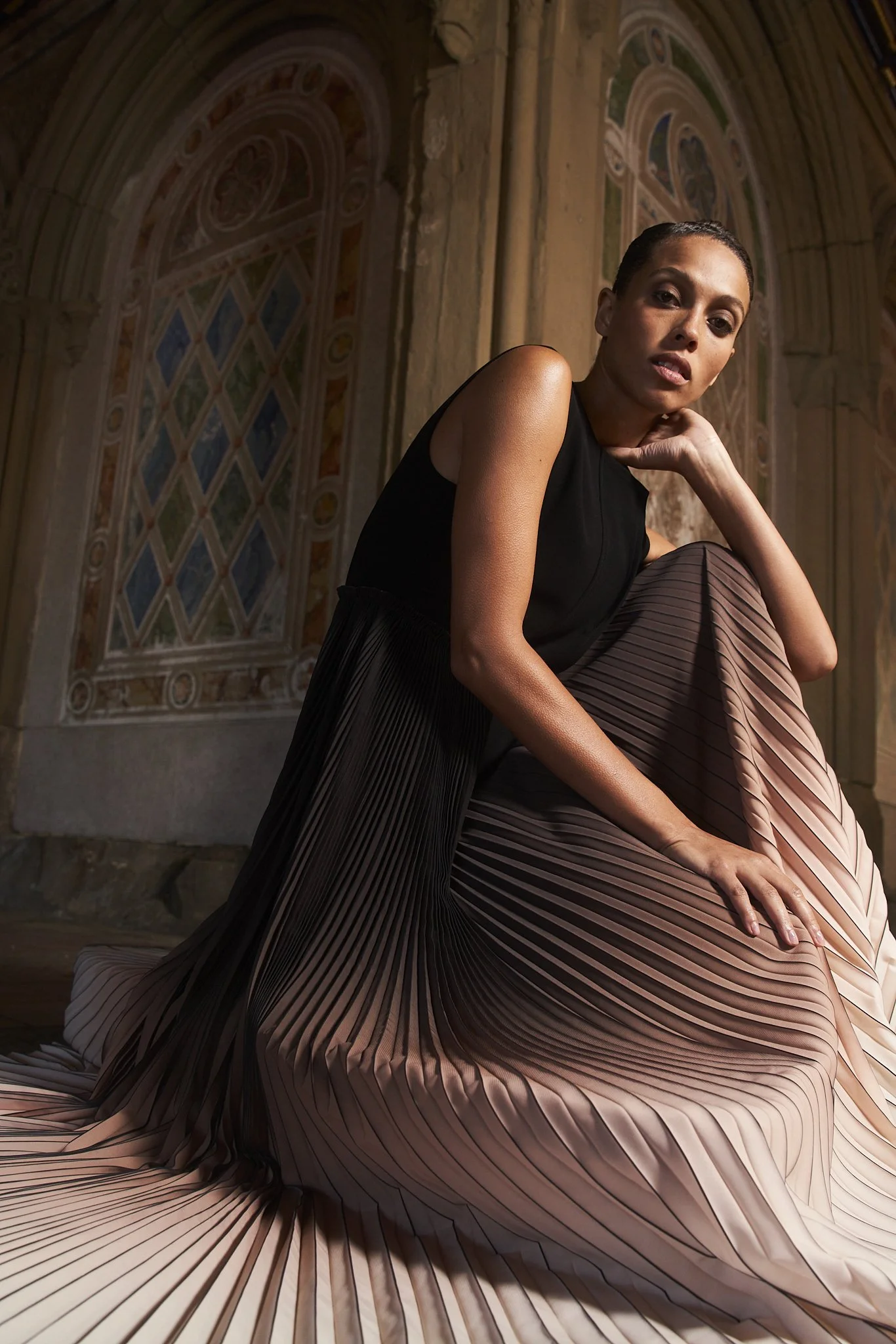 A woman with short hair and a black sleeveless top sits on pleated fabric in front of a decorated wall with large, arched stained glass windows.
