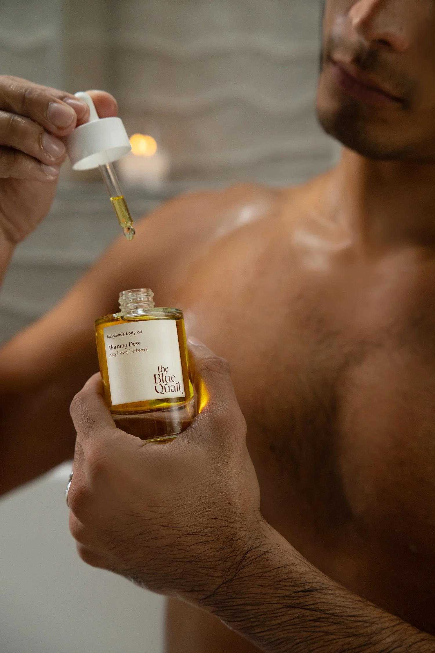 A shirtless man holding a bottle of yellow-colored body oil labeled 'Morning Dew' by The Blue Quail while a hand with a dropper is adding oil to his shoulder.