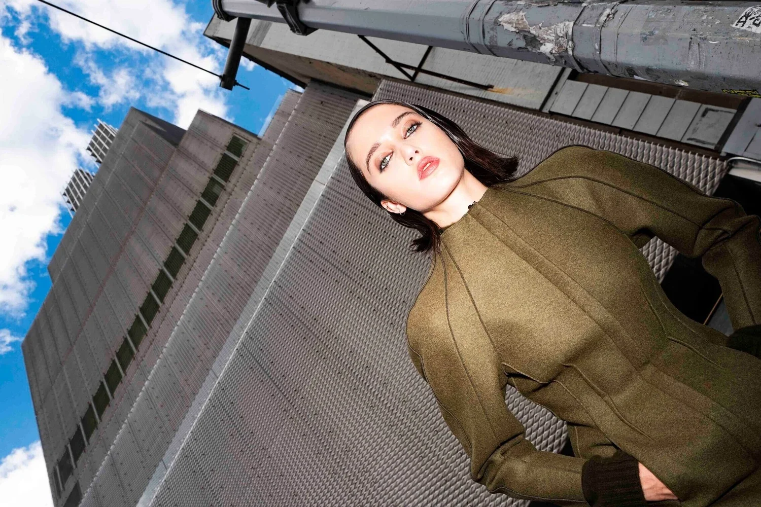 A woman with dark hair and light skin in an olive green jacket standing on a rooftop terrace with a cityscape in the background, including tall buildings and a blue sky with clouds.