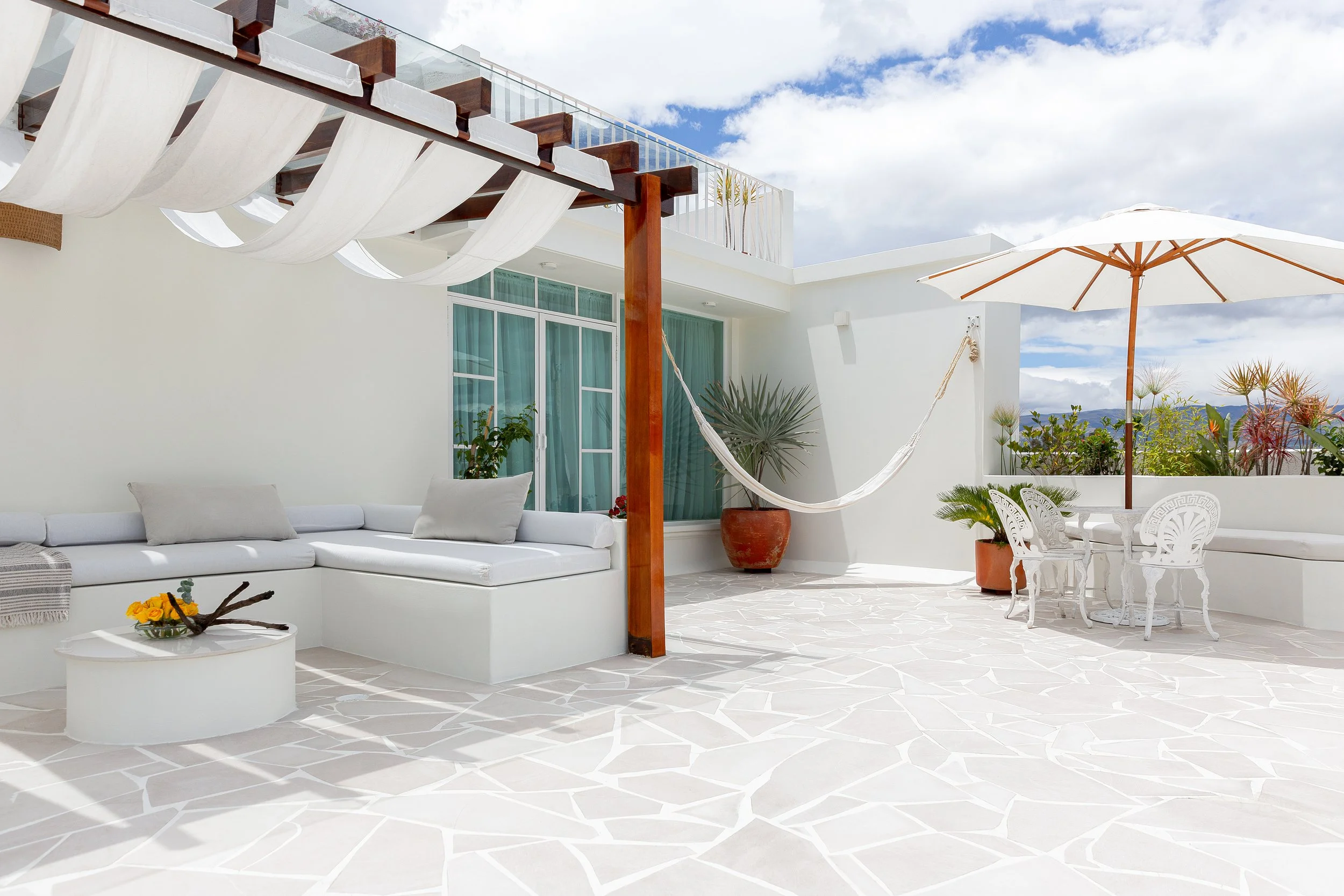 A spacious outdoor patio with white furnishings including a sectional sofa with white cushions, a round coffee table with a flower vase, and a white dining table with chairs. There is a large white umbrella, potted plants, and a hammock hanging from a wooden post. A balcony with glass railing and a turquoise curtain are visible, with a partly cloudy sky above.