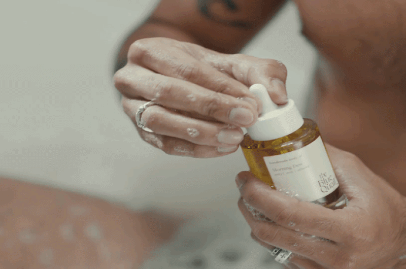 Person holding a brown glass bottle with a dropper cap, dispensing oil or serum onto their hand.
