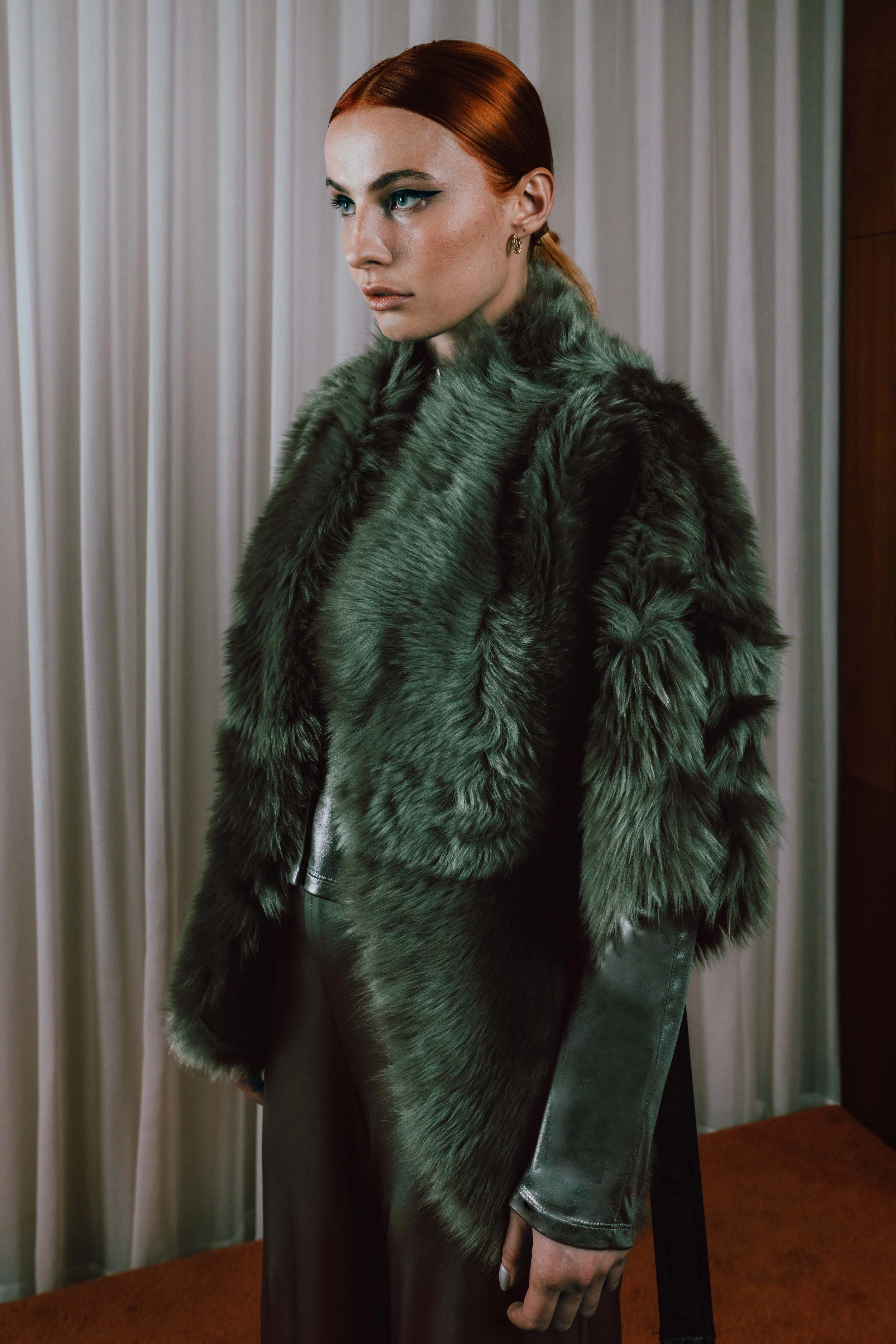 A woman with red hair and blue eyes wearing a dark green fur coat and black leather pants in front of a light-colored curtain.