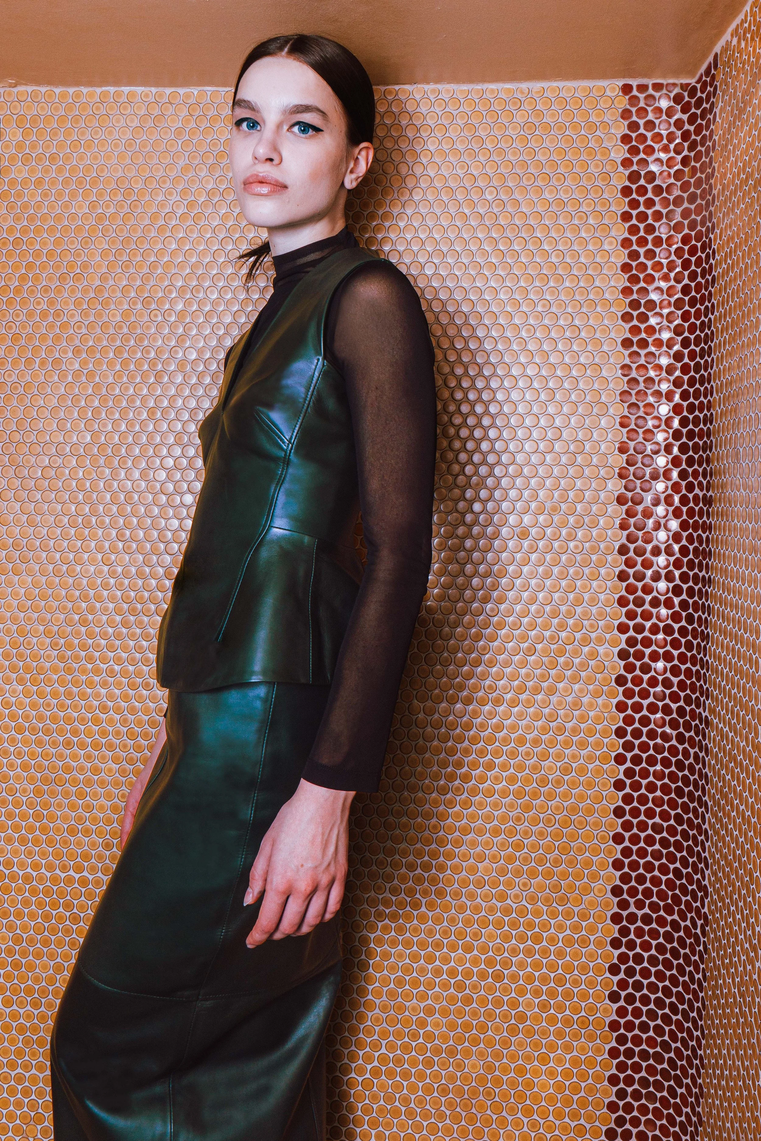 A woman with blue eyes and dark hair in a low ponytail standing against a hexagonal tiled wall wearing a black leather dress with sheer sleeves.