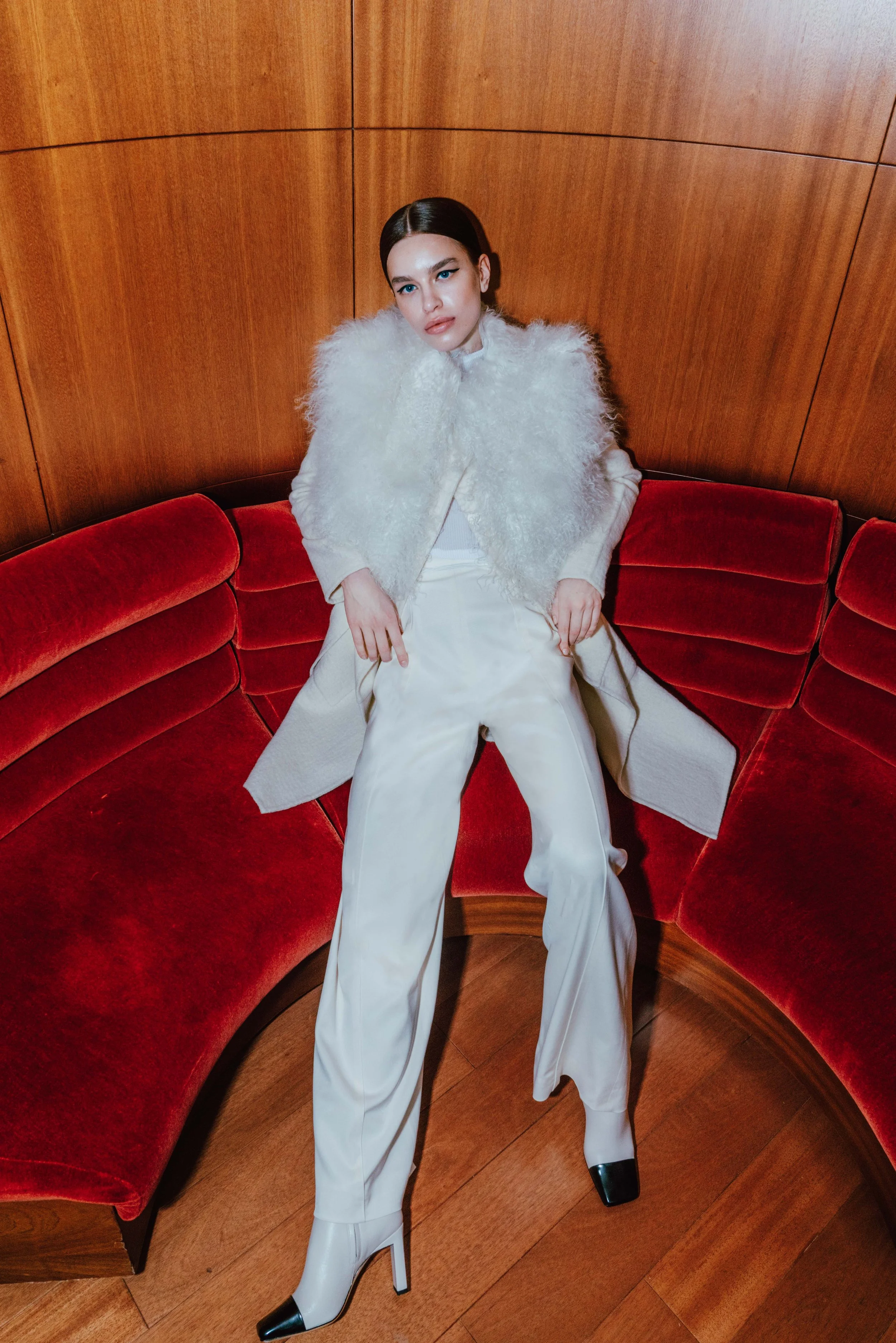 A woman with dark hair and light skin sitting on a red curved velvet sofa, wearing a white fluffy jacket, white wide-legged pants, and black patent leather high-heeled boots, with a wooden wall backdrop.