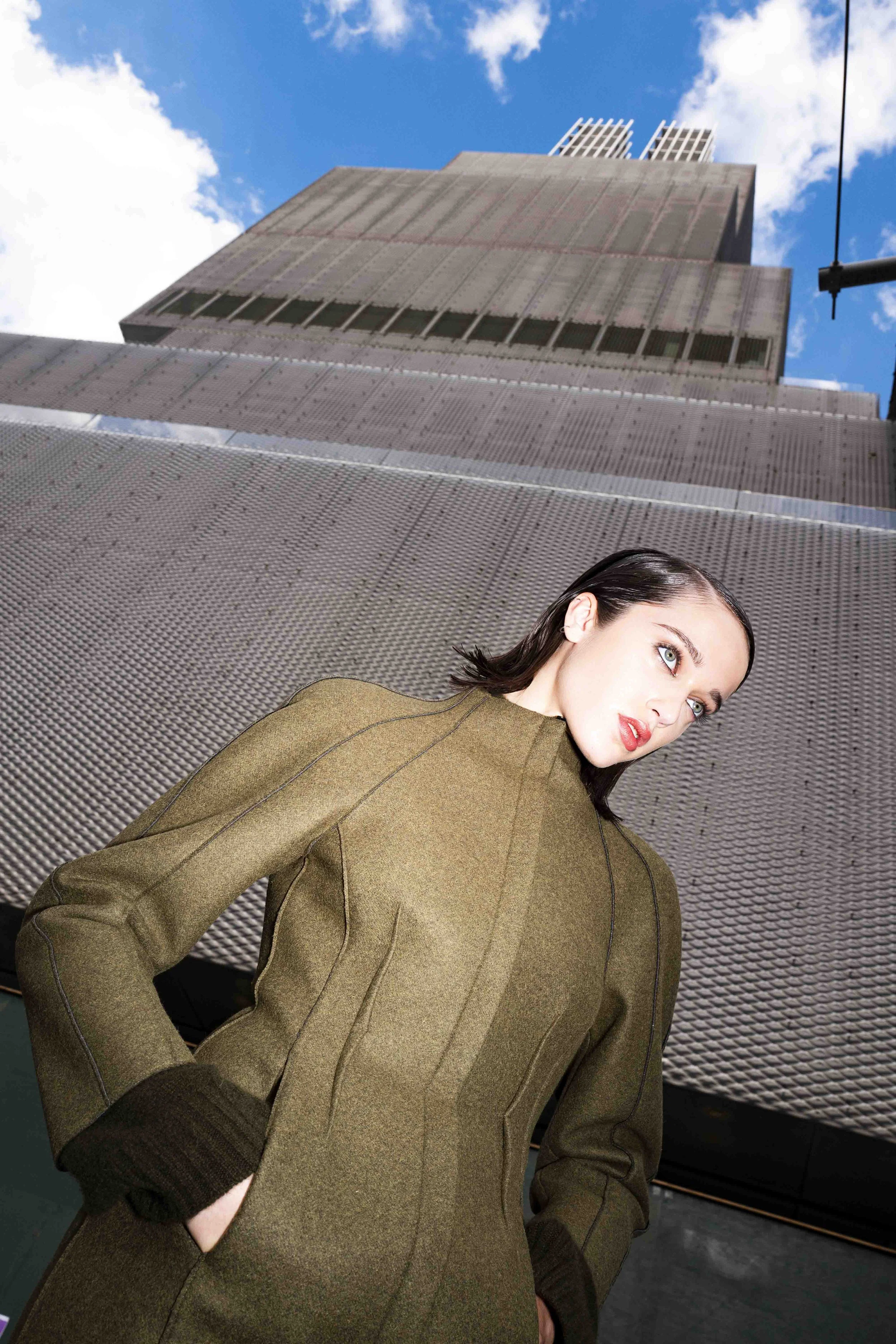 A young woman with dark hair and blue eyes wearing a khaki dress, standing outdoors on a metal surface with a modern building and a blue sky with clouds in the background.