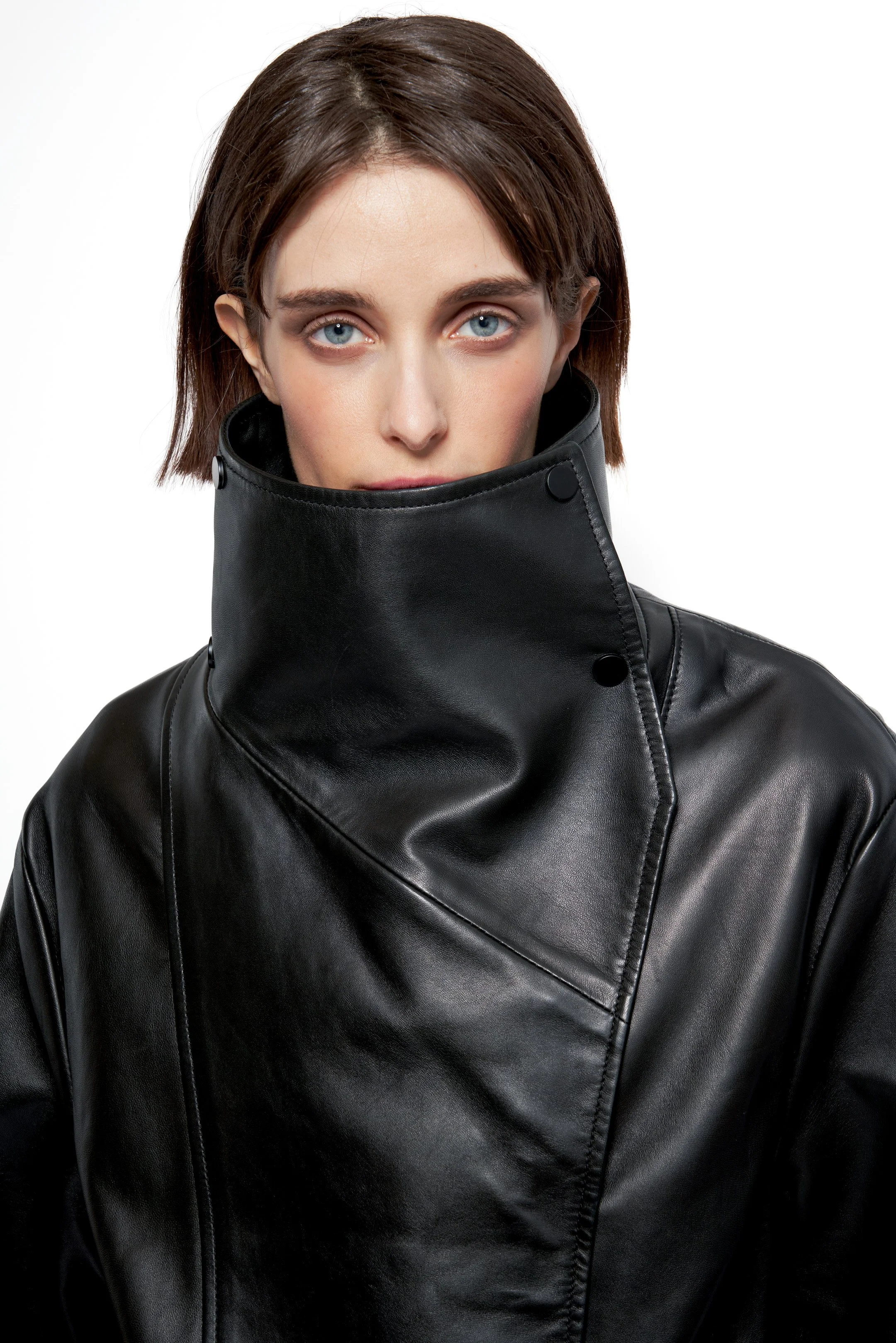 A woman with brown hair and blue eyes wearing a large black leather jacket with the collar up and buttons.