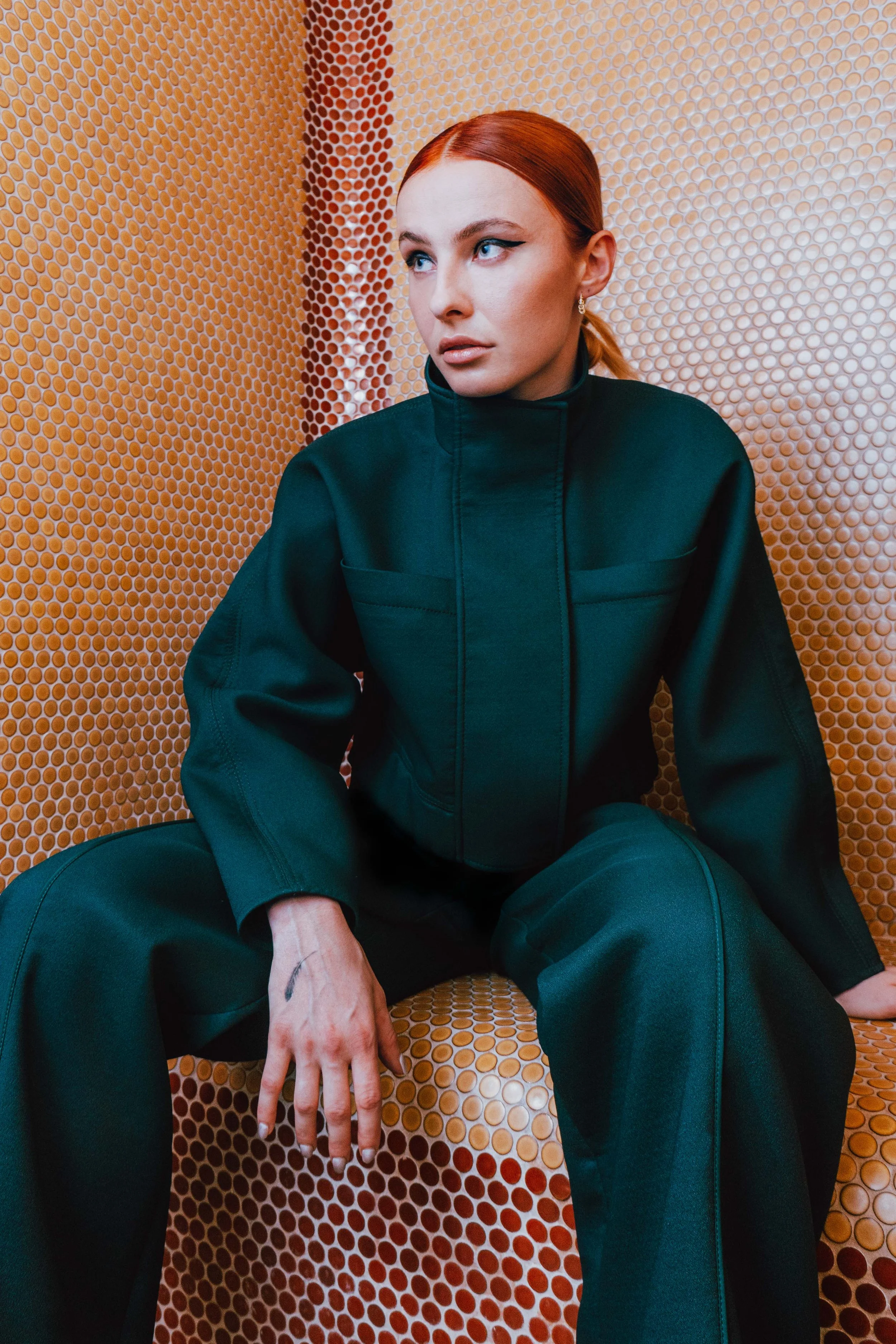 A woman with red hair, wearing a black outfit, sitting against a textured orange and red wall.