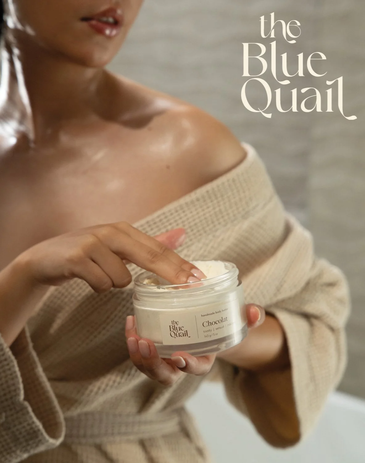 A woman in a beige sweater holding a jar of body butter labeled 'the Blue Quail' and 'Chocolat,' with her finger dipping into the product.