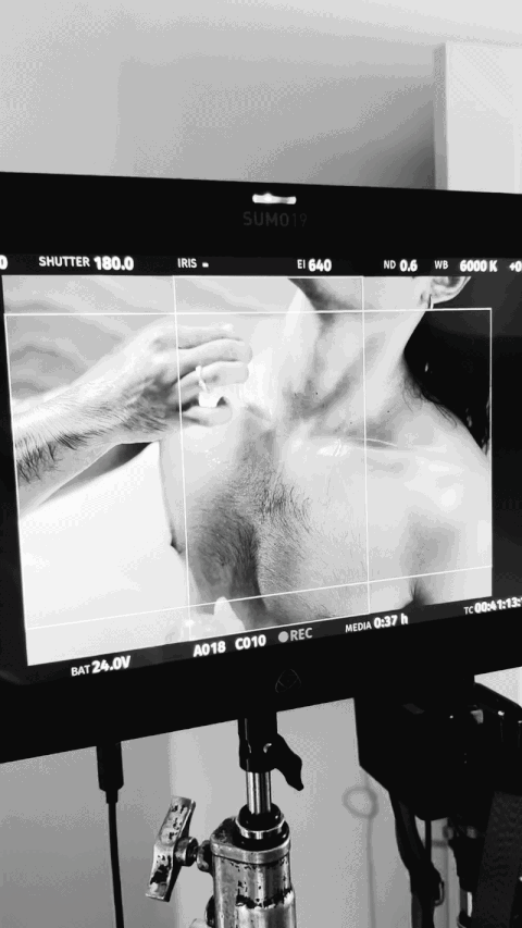 Black and white photo of a camera monitor showing a close-up of a person's chest and hand, with the person adjusting their collar.