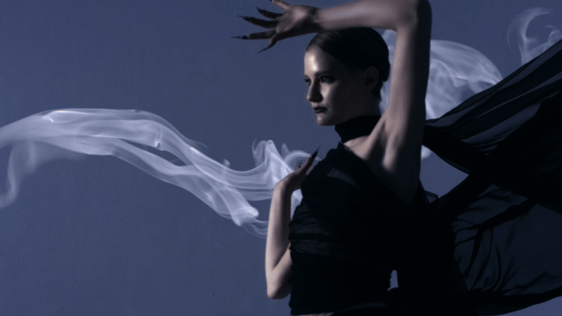 A woman in black clothing poses with her arms raised and flowing fabric around her against a dark background.
