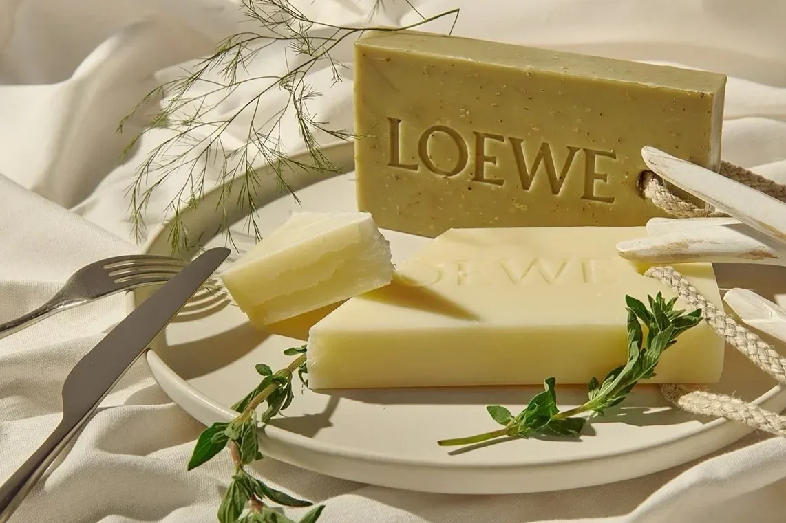Three types of soap with the brand name 'LOEWE' embossed on two of them, arranged on a white plate with green herbs, a fork, and a knife, on a white cloth background with decorative foliage.