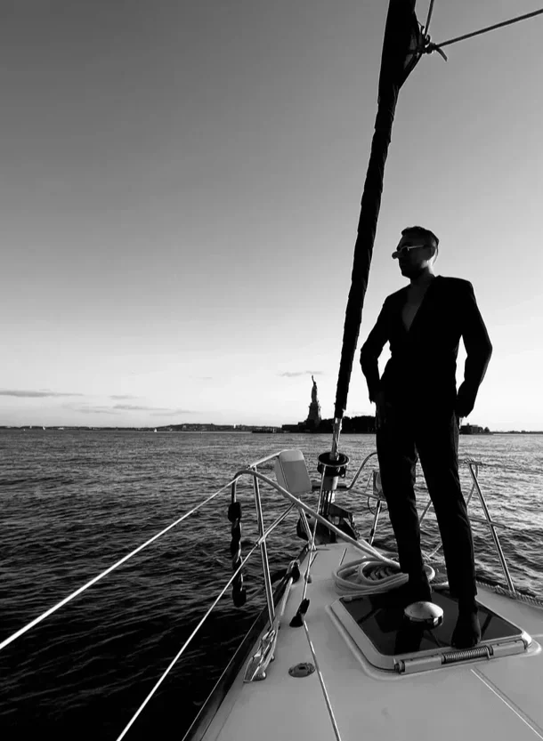 A man in a suit and sunglasses standing on a sailboat at sunset, with the Statue of Liberty visible in the background.