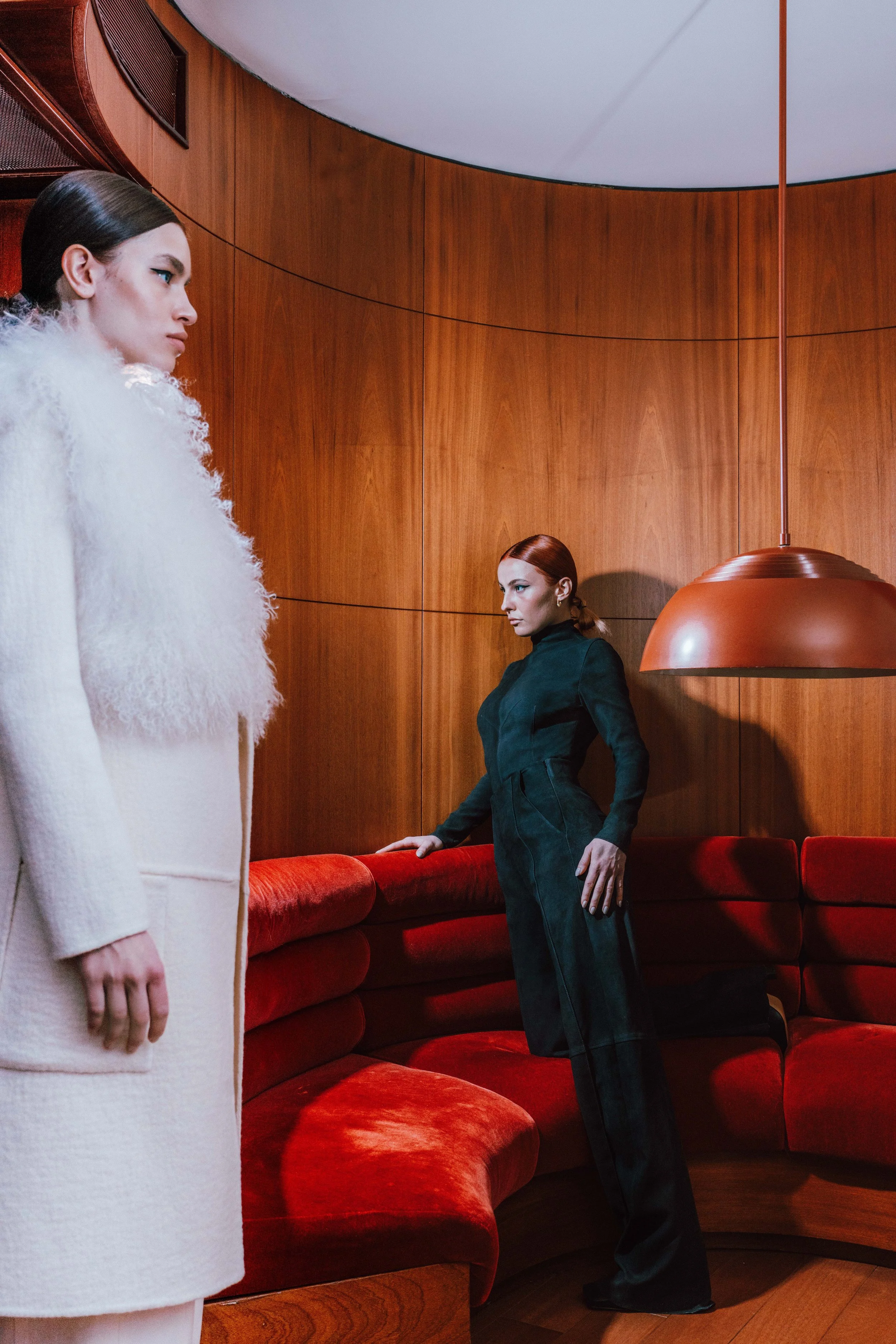 Two women in a modern, wood-paneled room with red velvet seating and a large copper pendant light. One woman with dark hair in a white, textured coat stands to the left, partially in frame. The other woman with red hair in a dark suit leans on the red velvet sofa, looking towards the woman in white.